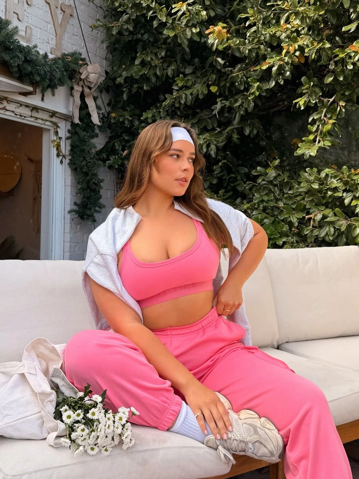 Woman sitting on a white couch wearing a pink sports bra and matching pink sweatpants, with a white headband and a white open shirt, outdoors near green bushes and a wall with festive decorations.