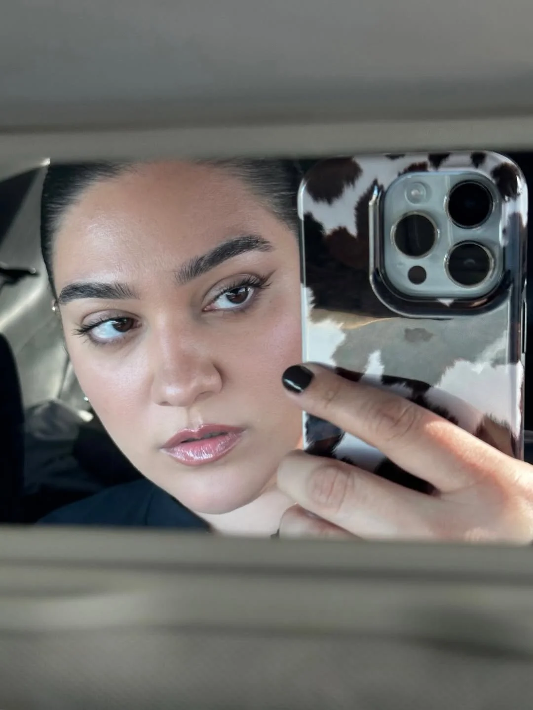 Close-up of a woman with short dark hair and light makeup taking a selfie in a mirror with her phone, which has a cow print case.