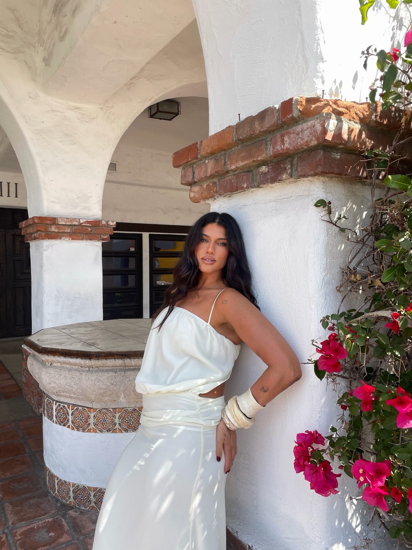 A woman with long dark hair wearing a white dress and bracelet, standing outdoors next to a white stucco wall with a cactus and pink flowers, under an arched patio with brick accents.