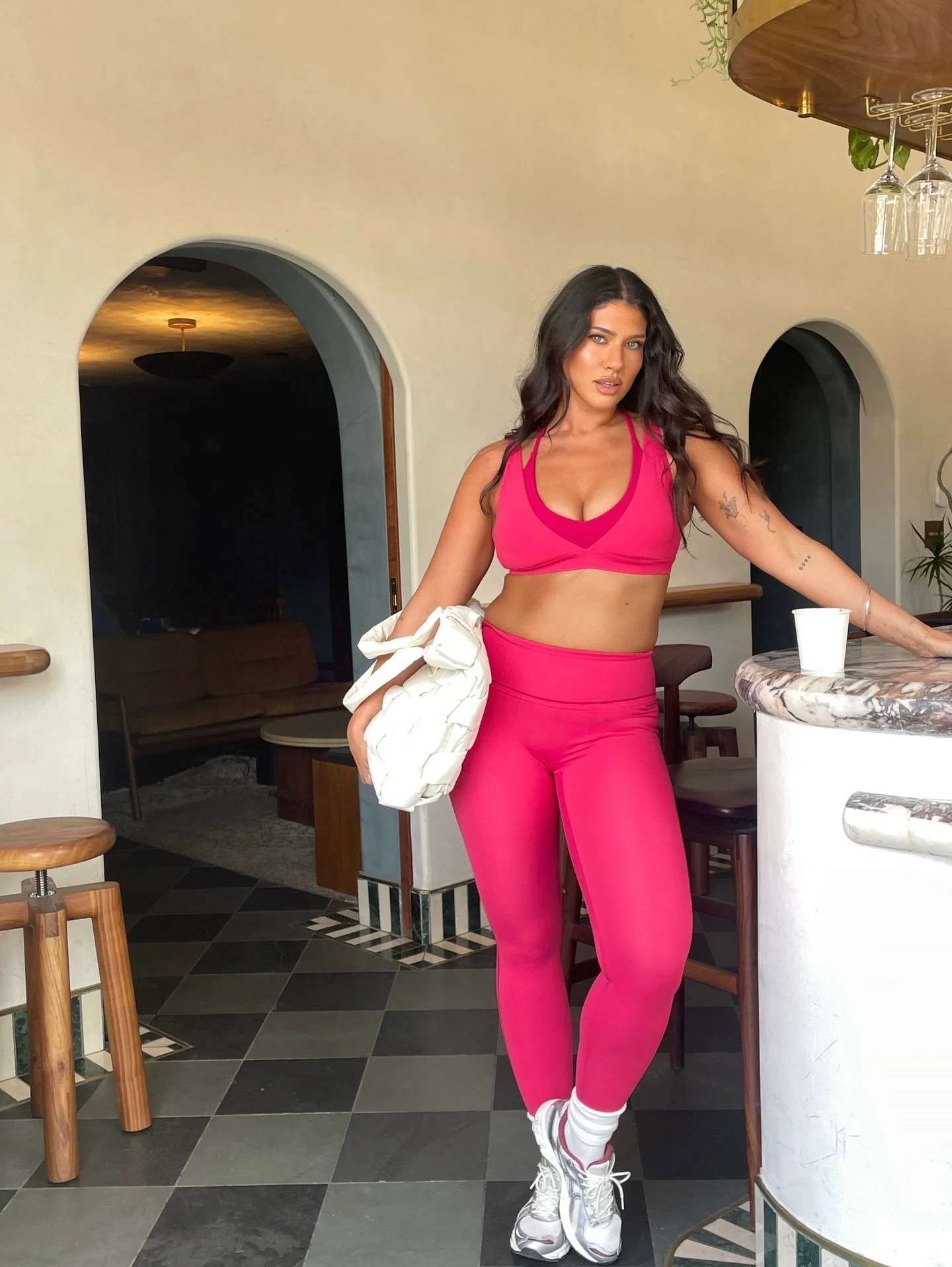 A woman with long dark hair and tattoos wearing a pink sports bra and leggings, standing at a kitchen counter with a cup, in a modern, stylish room with a marble countertop, wooden chairs, and arched doorways.