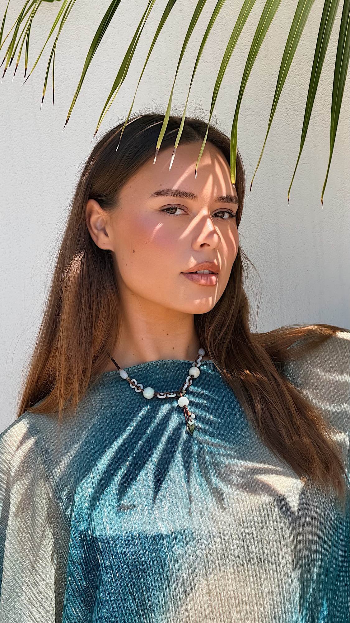 A woman with long brown hair, wearing a beaded necklace, stands in front of a white wall with palm leaf shadows, and a large palm leaf shading part of her face.