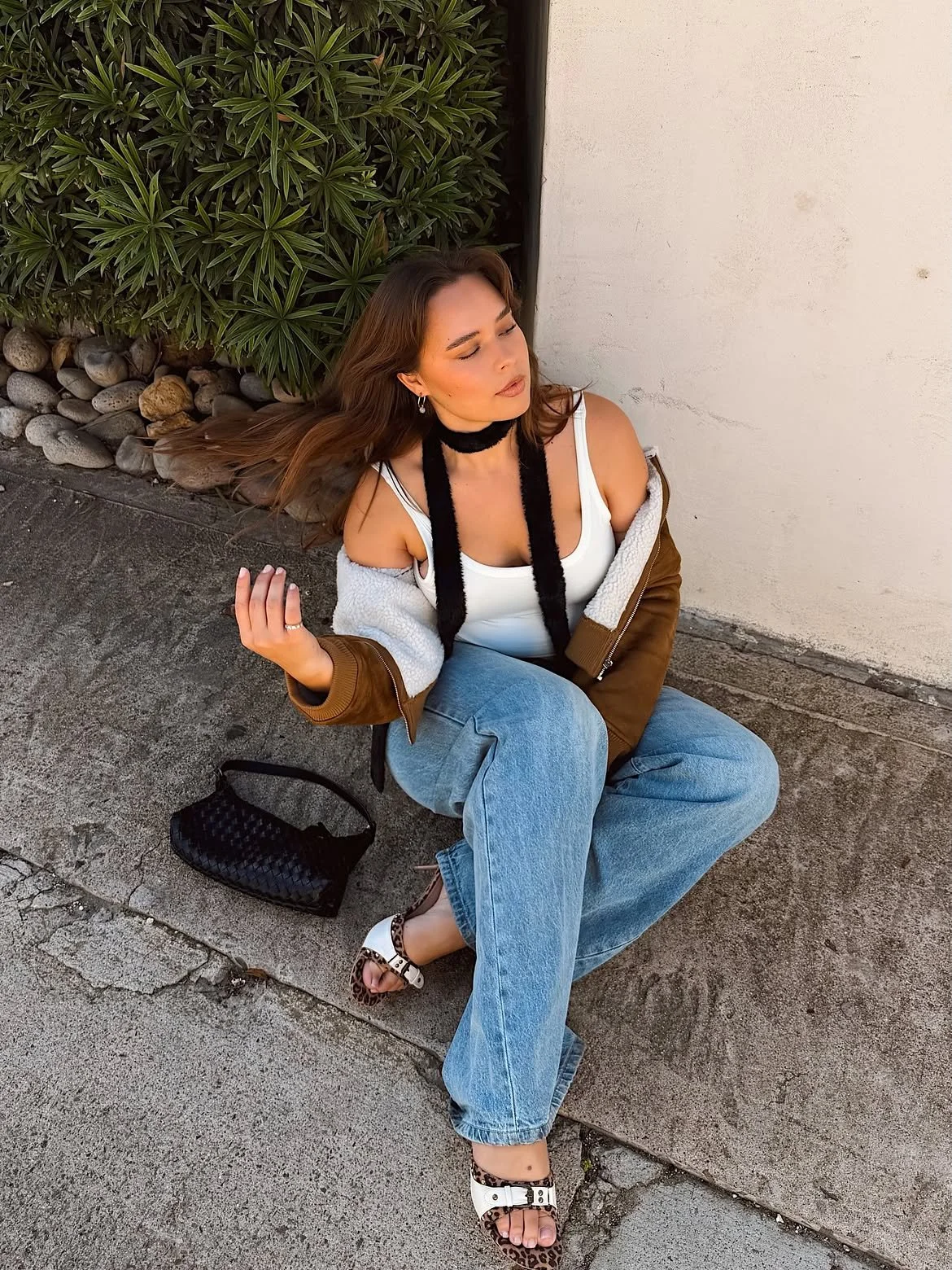 A woman with long brown hair sitting on the sidewalk near a white wall and green bushes, wearing a white tank top, a brown jacket with white fleece lining, wide-leg jeans, and white sandals with animal print details.