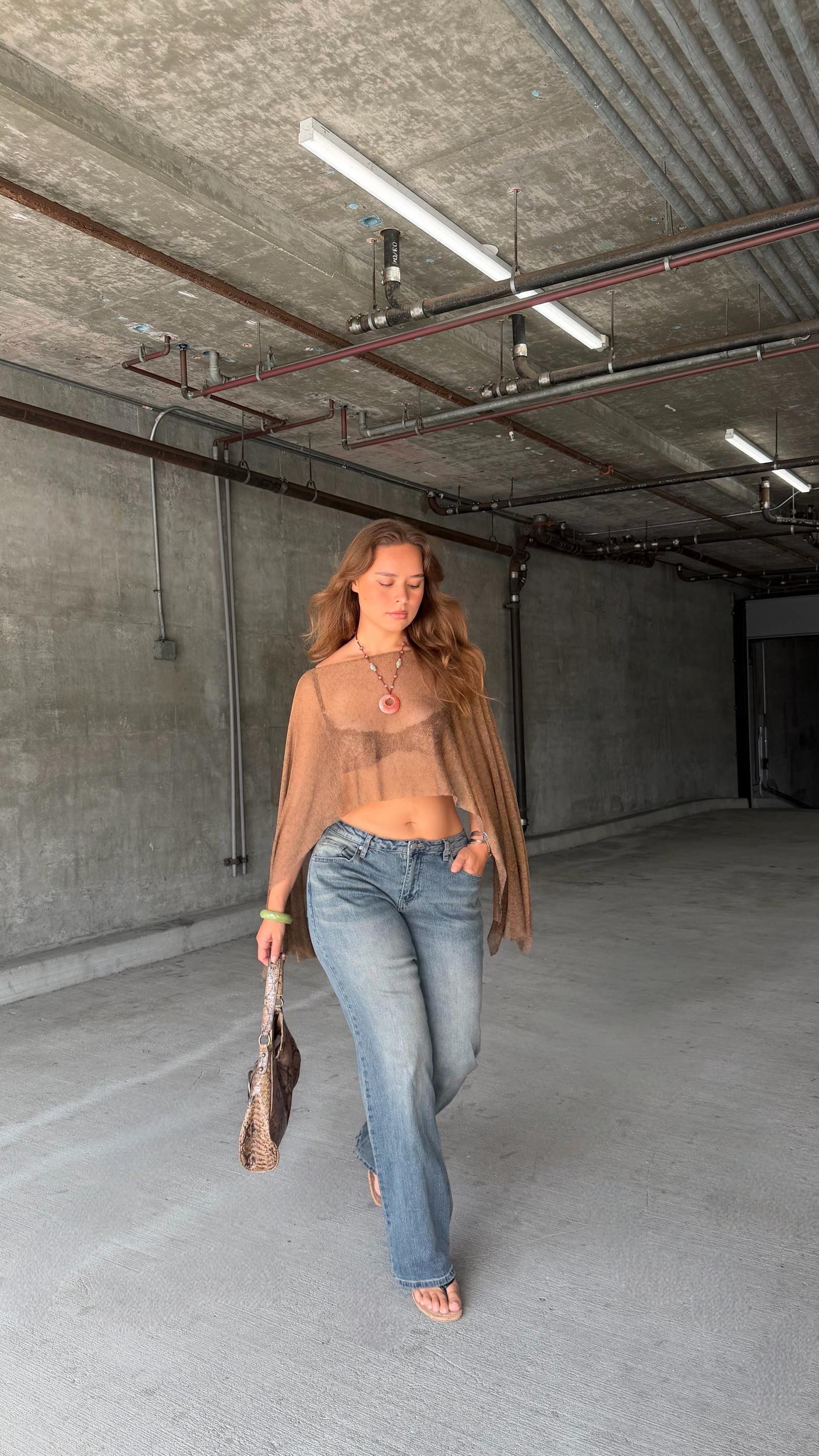 A young woman walking in an empty parking garage with concrete walls and ceiling, wearing a sheer brown top, blue jeans, and sandals, carrying a snake-print handbag.