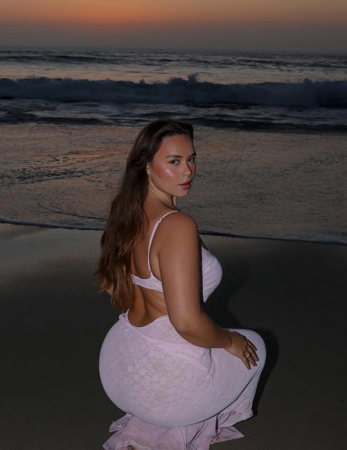 A woman with long brown hair kneeling on the beach at sunset, wearing a pink dress, with the ocean waves in the background.
