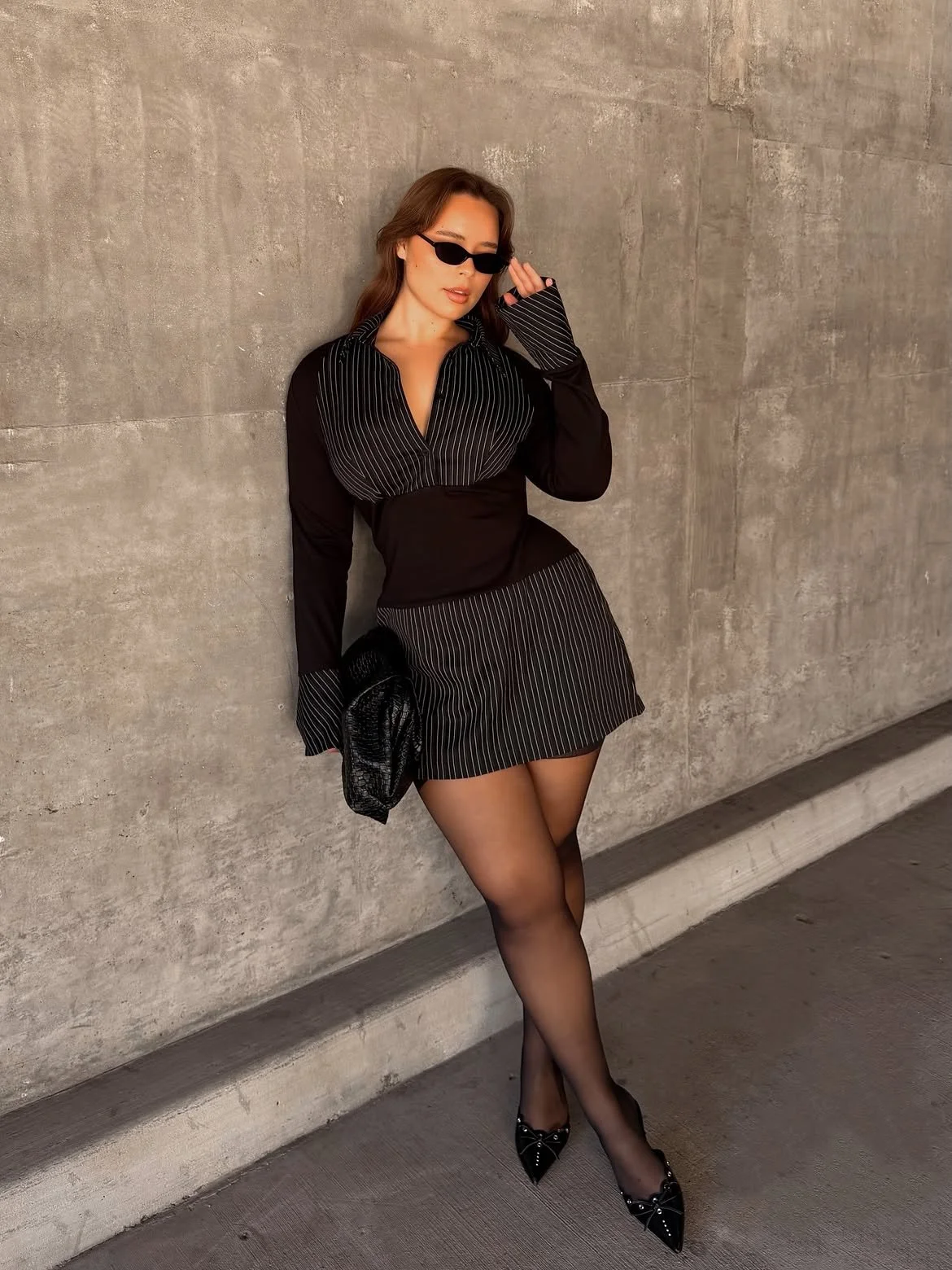 A stylish woman wearing a black pinstripe dress, black stockings, and high heels, posing against a concrete wall with sunglasses and a small clutch purse.