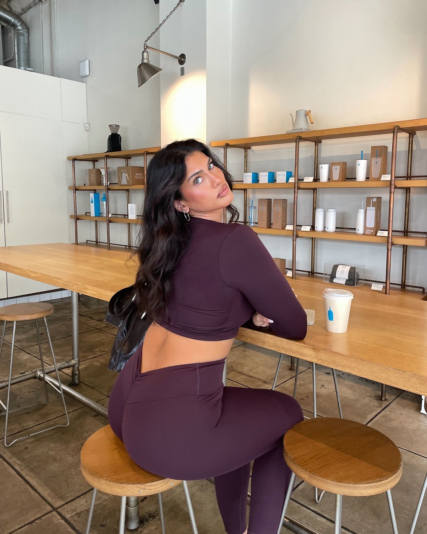 A woman with long dark hair and earrings sitting at a wooden table inside a cafe, wearing a dark purple sports top and matching leggings, with a coffee cup in front of her and shelves with various cups and products in the background.