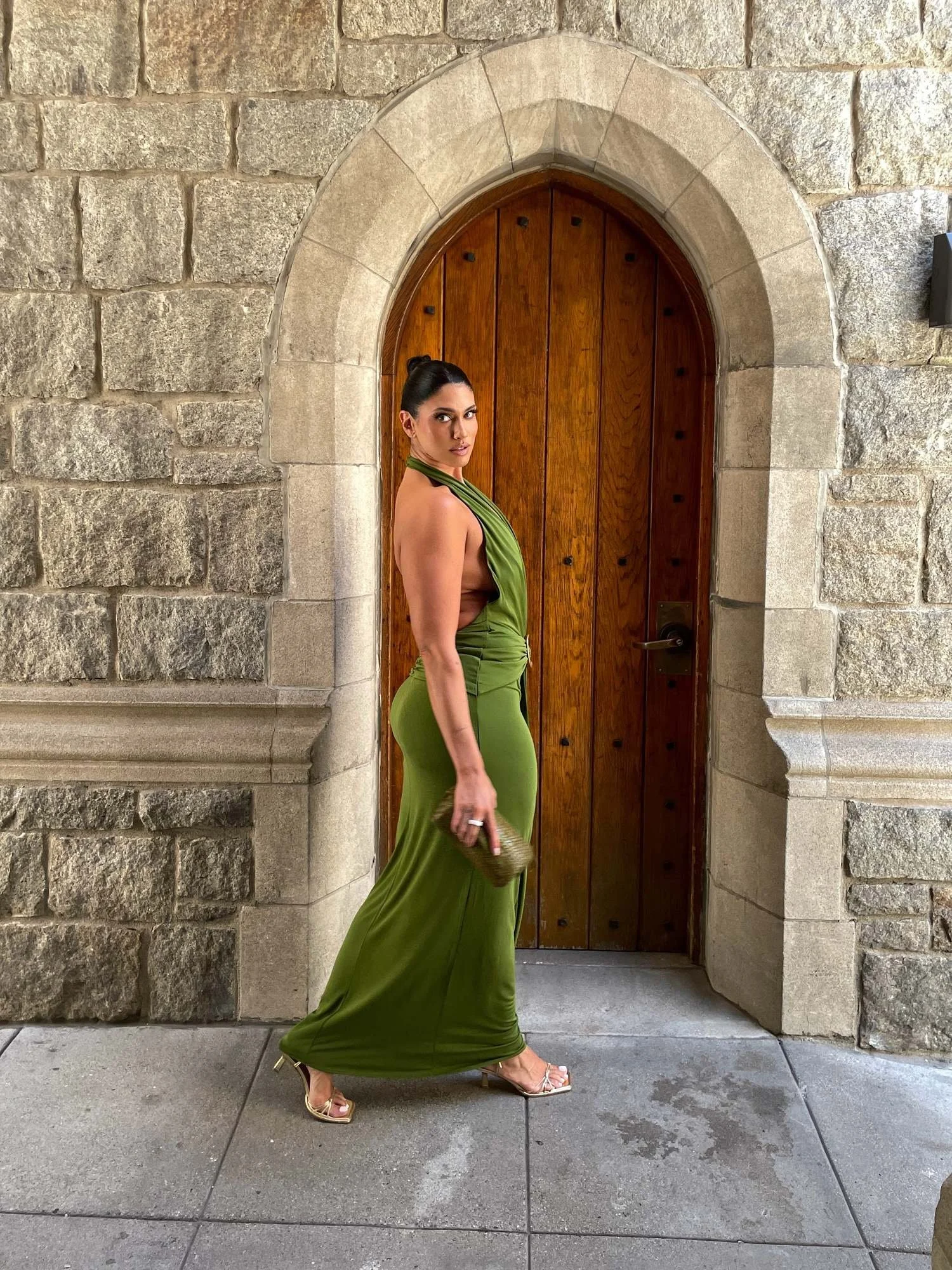 A woman in an elegant green satin dress, heels, and gold clutch purse stands in front of a wooden arched door framed by stone wall.