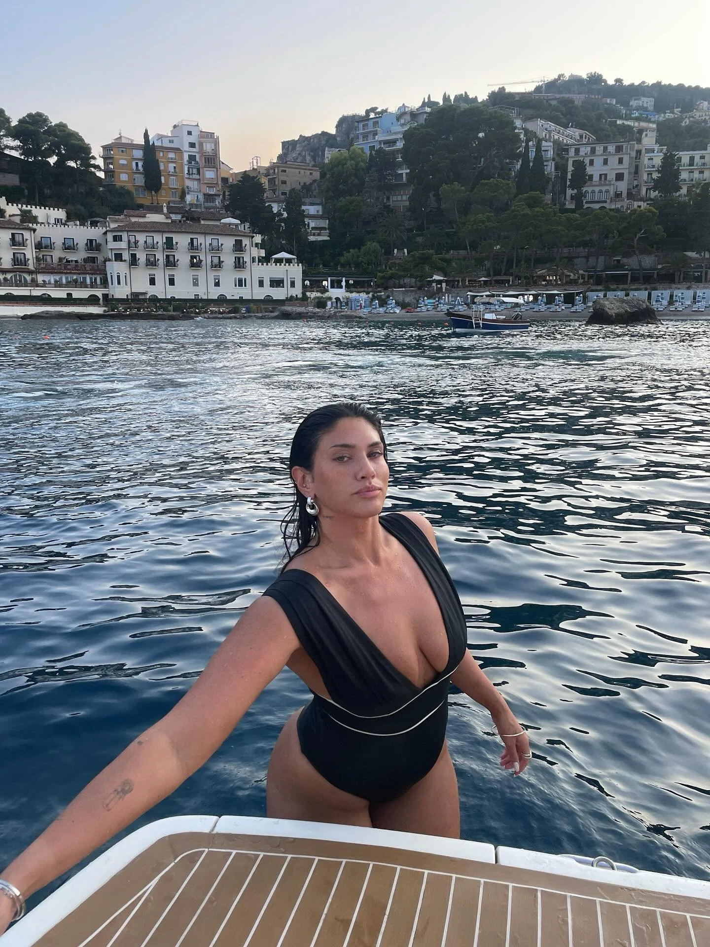 Woman in a black swimsuit taking a selfie on a boat, with water and buildings on a hillside in the background.