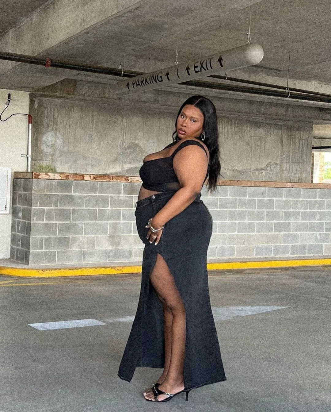 A woman with long black hair stands in a parking garage wearing a black crop top and a long black skirt with a high slit, black high-heeled sandals, and jewelry.