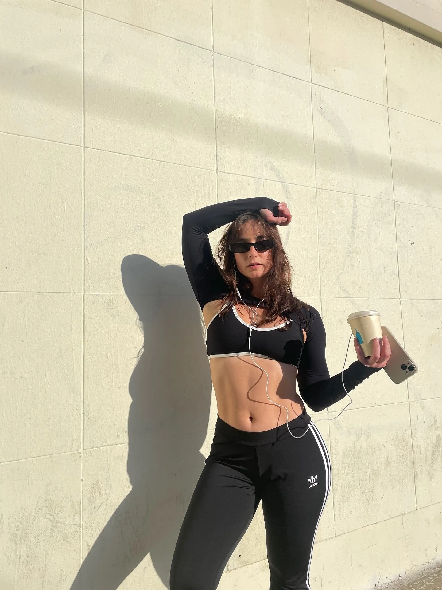 A woman in black activewear, sunglasses, and headphones, holding a phone and coffee cup, standing against a beige tiled wall in sunlight.