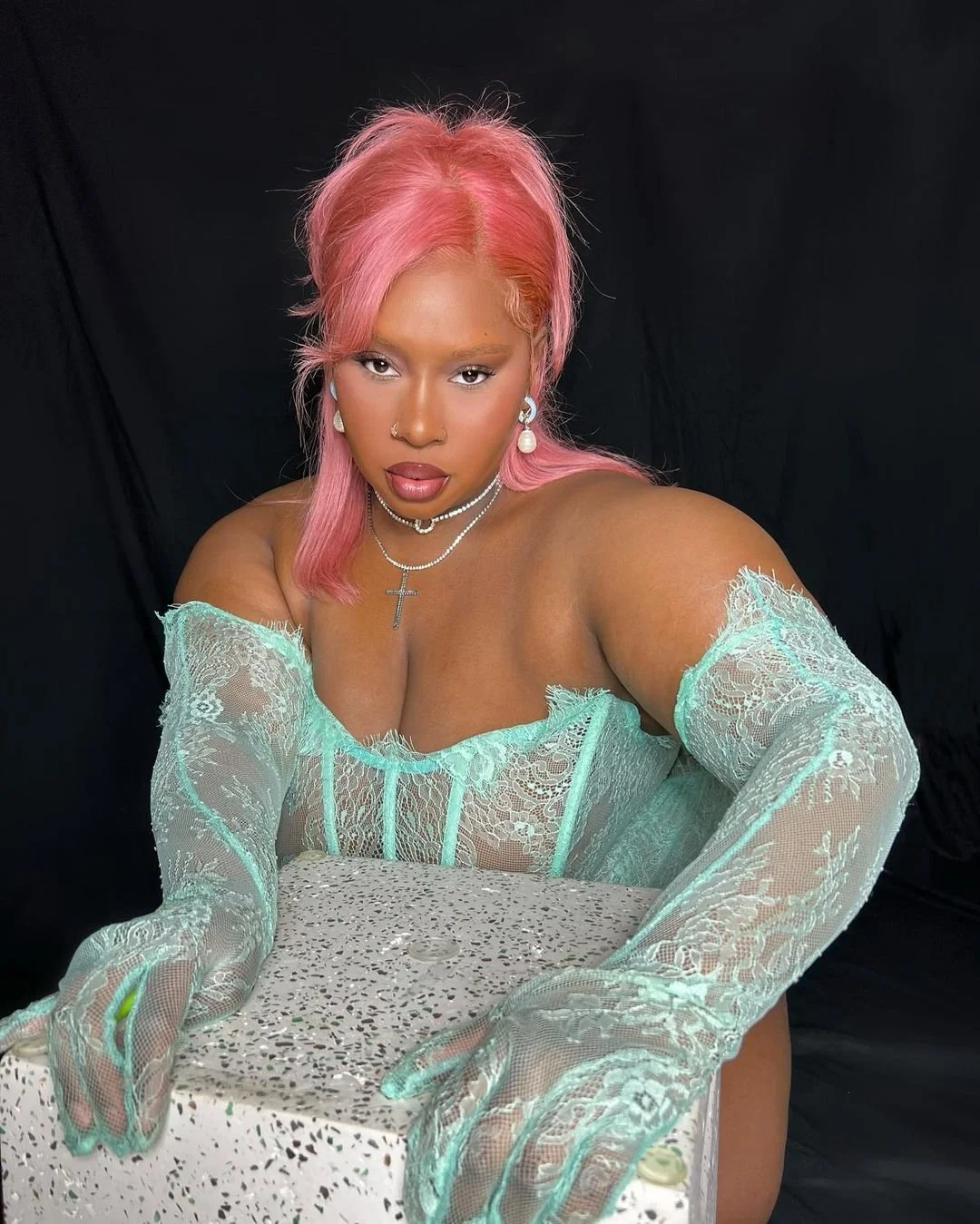 Woman with pink hair wearing lace gloves and jewelry, posed with a glittery box against a black background.
