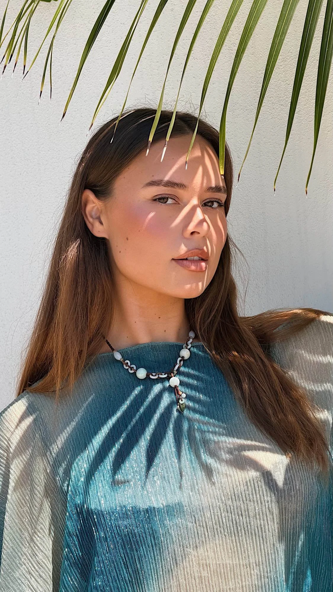 A woman with long brown hair stands in front of a white wall, with shadows cast by palm leaves across her face and clothing. She is wearing a blue and cream satin-like blouse and a necklace with white beads and a small pendant.