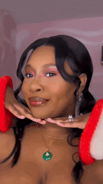A woman with dark hair styled in loose curls, wearing pink eye makeup and red lipstick, is posing with her head resting on her hands, which are decorated with red and white furry cuffs. She is wearing a green pendant necklace and has a confident, joy