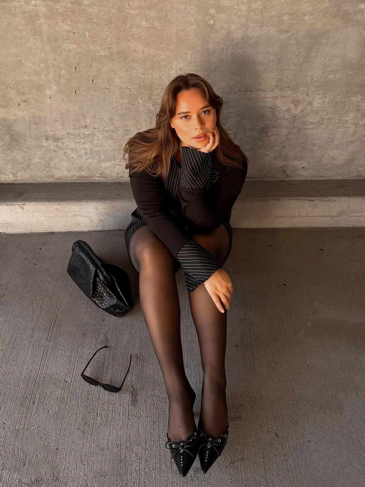 A woman sitting on the ground against a concrete wall, wearing black sheer tights, a black pinstripe blazer, black pointed heels, sunglasses, and a black textured handbag nearby.