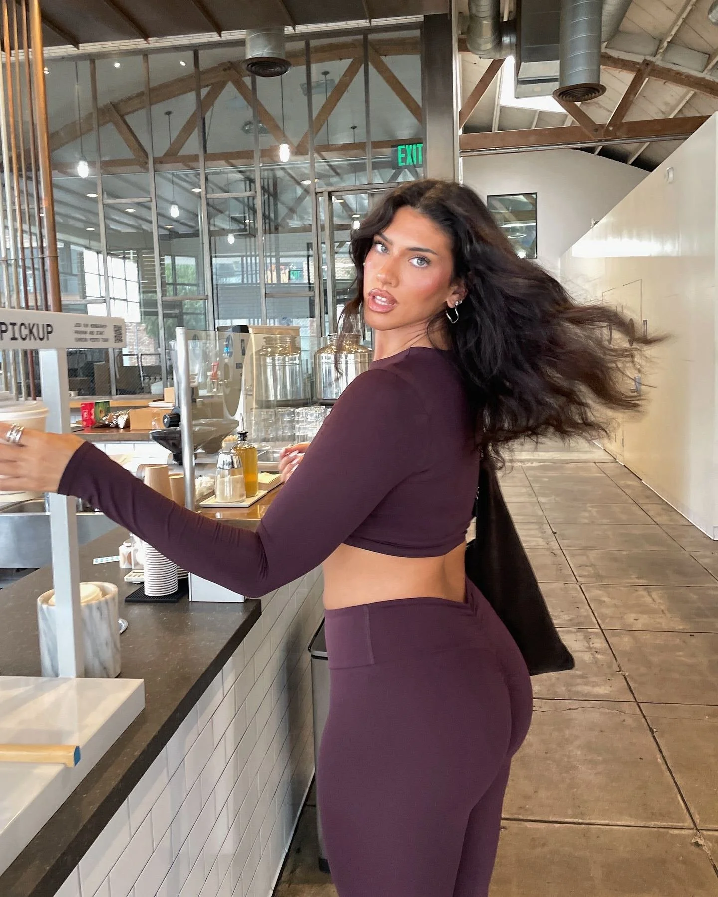 A woman with long dark hair and hoop earrings, dressed in a matching maroon long sleeve crop top and high-waisted pants, is at a counter in a modern restaurant or cafe, possibly ordering or preparing a drink.