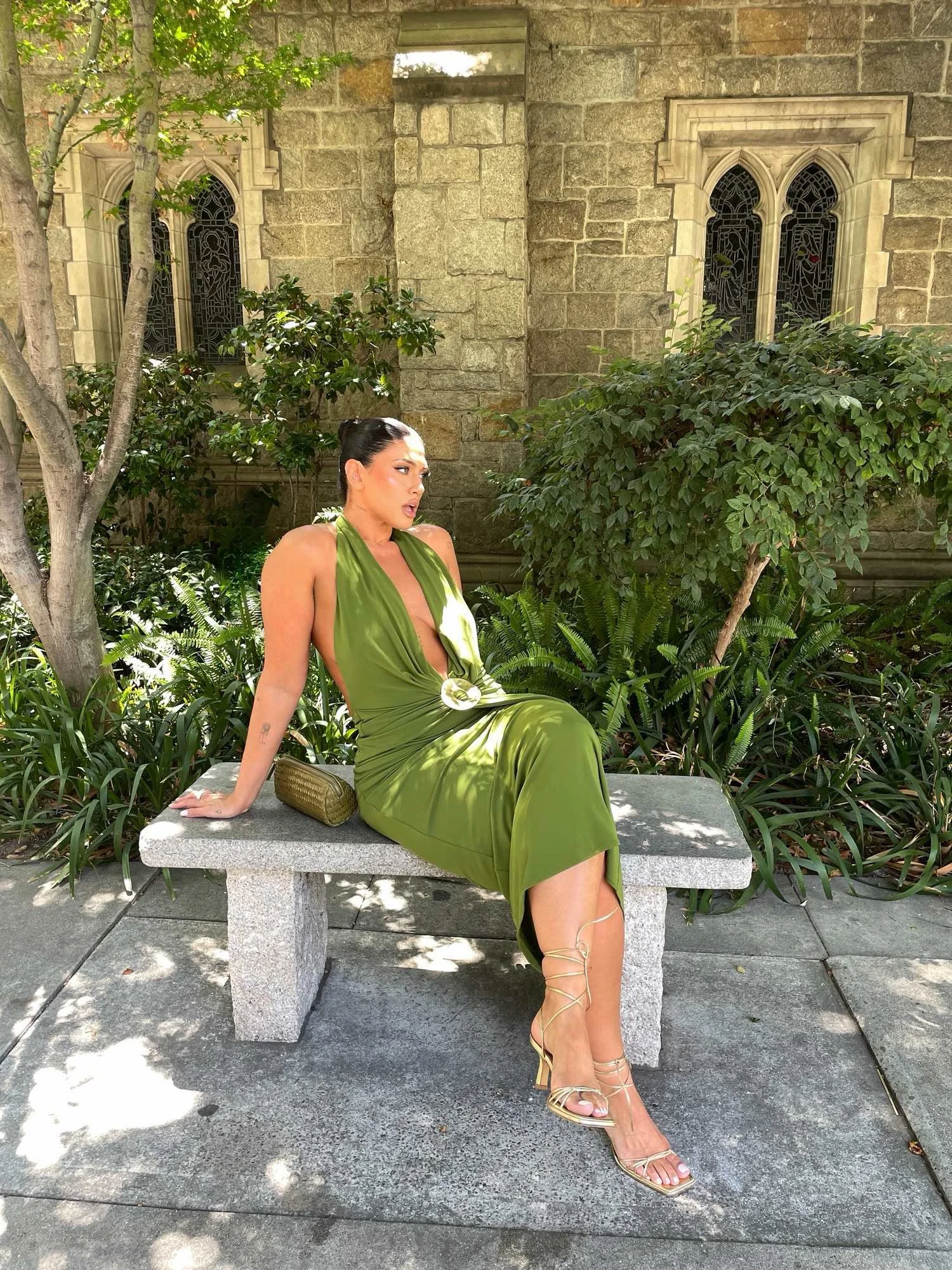 Woman in a green dress sitting on a stone bench outdoors, surrounded by trees and foliage, with a stone building and stained glass windows in the background.