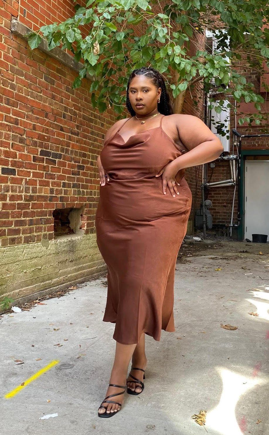 A woman in a brown satin dress standing outdoors against a brick wall with greenery, wearing black strappy heels and jewelry.