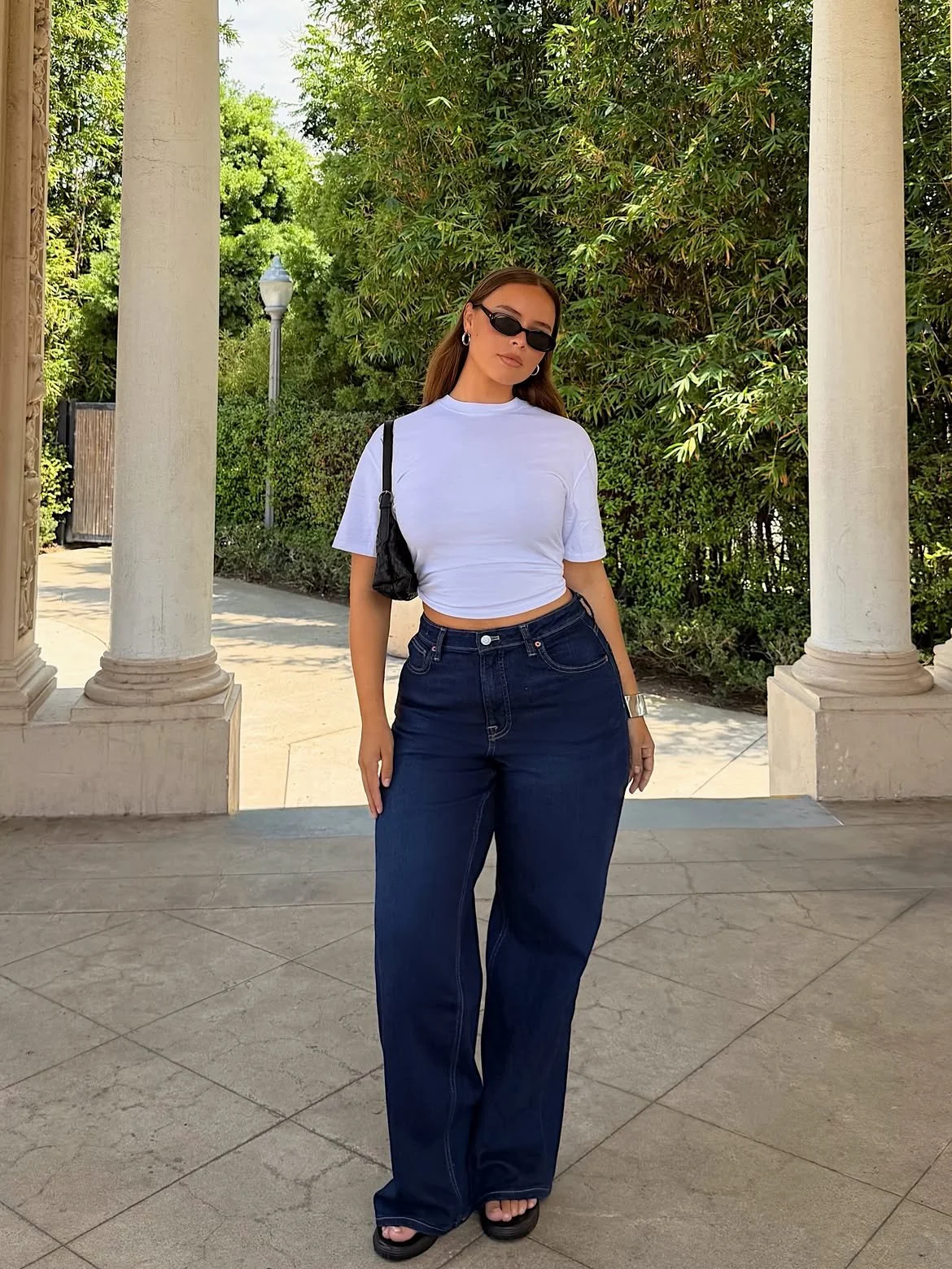 Woman standing outdoors near columns, wearing sunglasses, a white t-shirt, high-waisted jeans, and carrying a black purse.