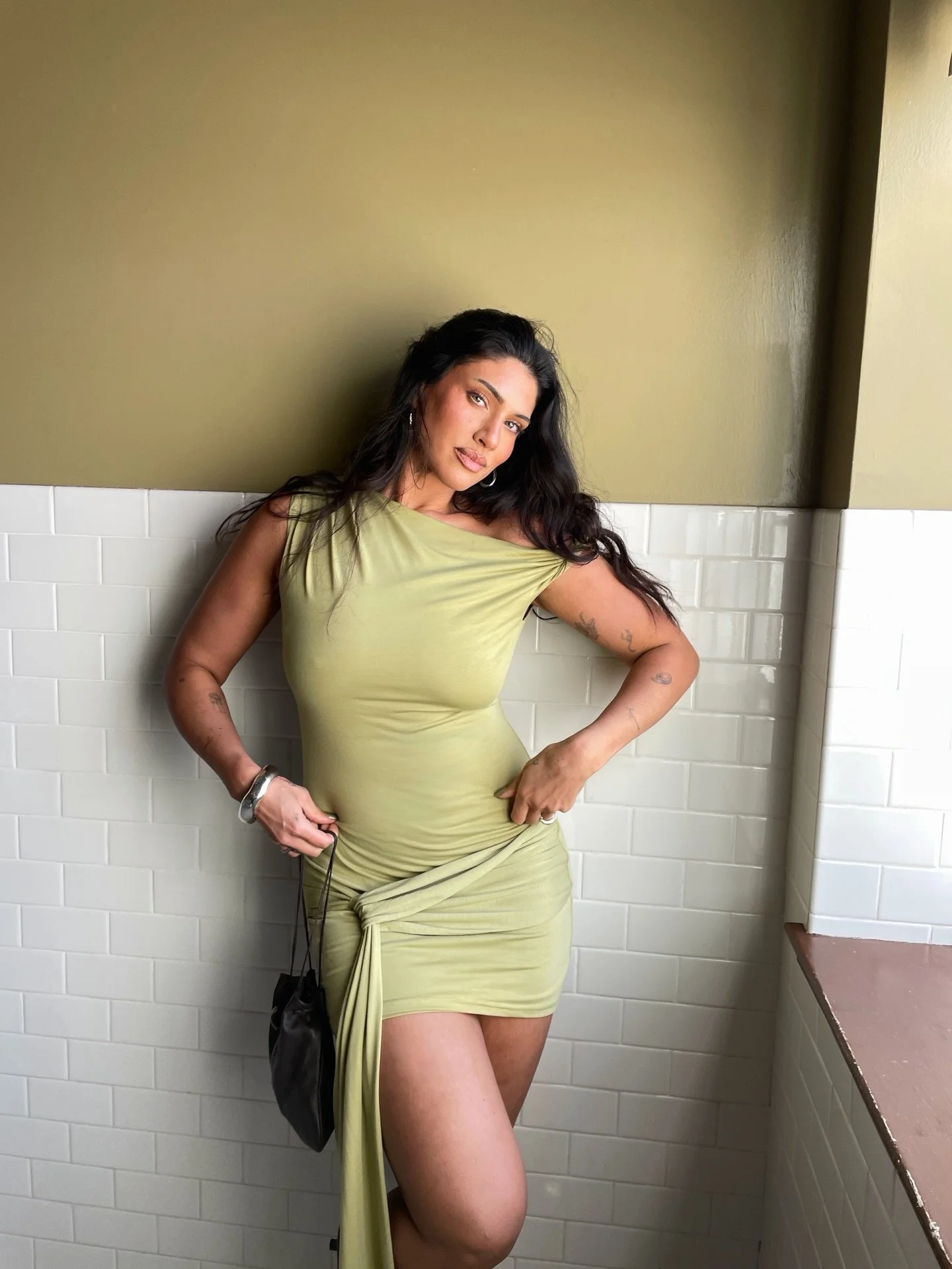 A woman with dark hair, wearing a light green dress, poses against a wall with white tiles and a green wall. She has tattoos on her arms and is holding the fabric of her dress while looking at the camera.