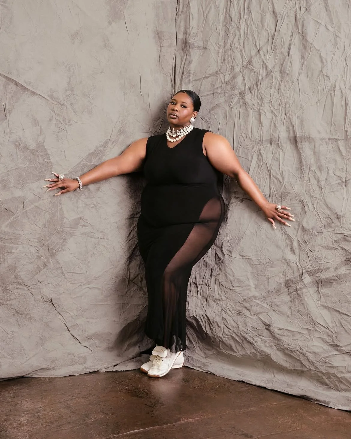 A woman wearing a black dress with sheer panels, accessorized with pearl jewelry, white sneakers, and rings, posing against a crumpled gray backdrop.
