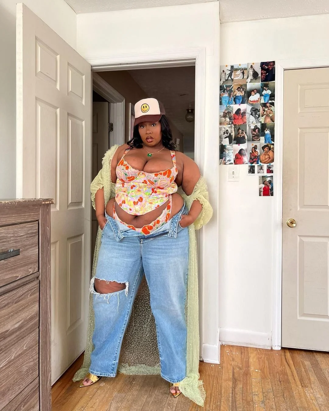 A woman standing in a room in front of a white wall with a collage of photos, wearing a floral top, ripped jeans, a green necklace, and a cap with a smiley face, posing with her hands in her pockets.