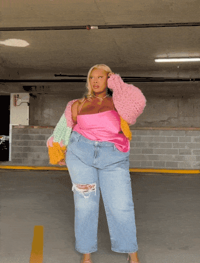 A woman posing in a parking garage, wearing a bright pink and yellow top with a pink fuzzy cardigan and ripped jeans.