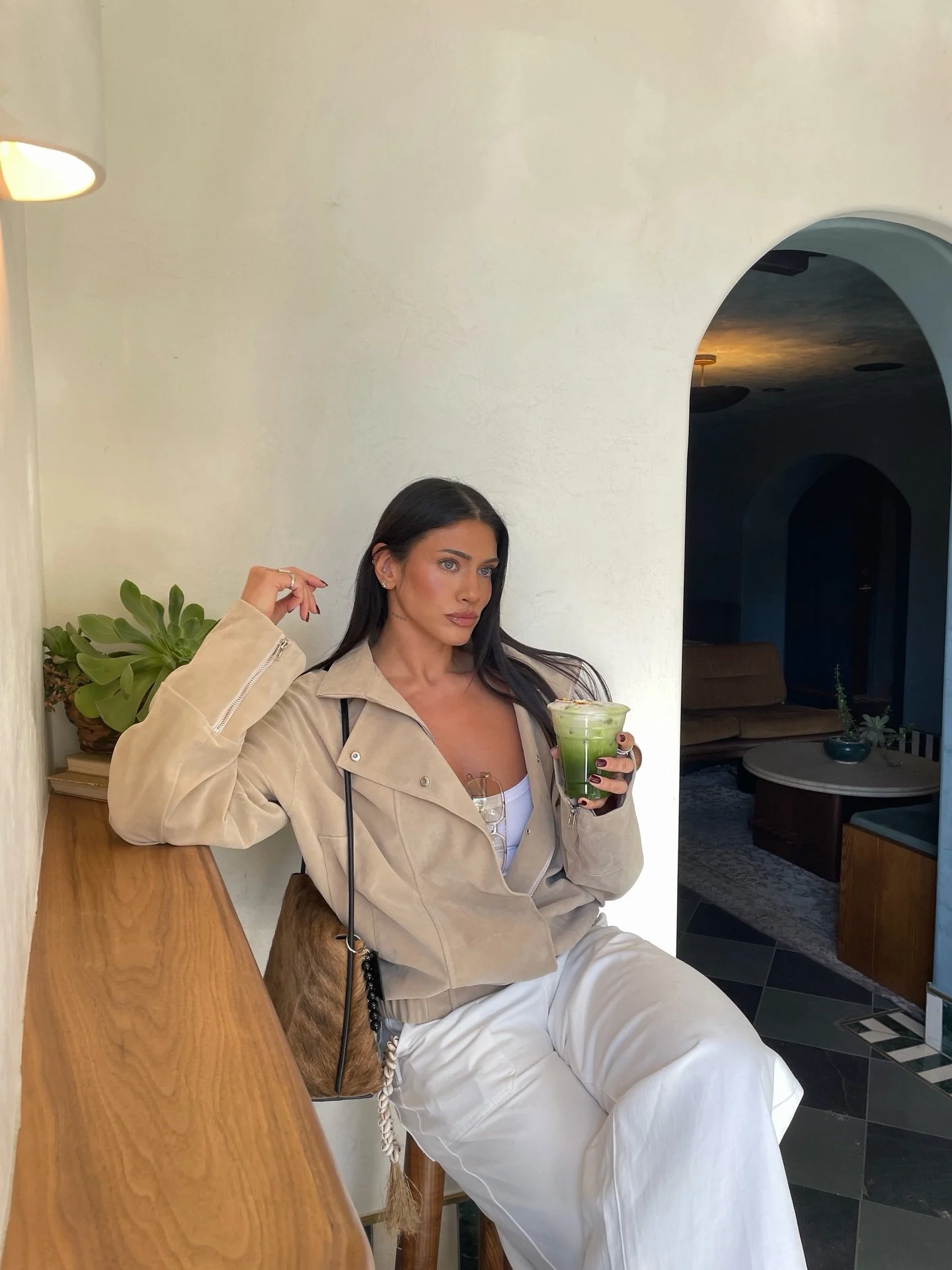 A woman with dark hair and light skin sitting at a wooden counter in a cafe, holding a green beverage, wearing a beige jacket and white pants, with a plant in the background and an arched doorway leading to a cozy lounging area.