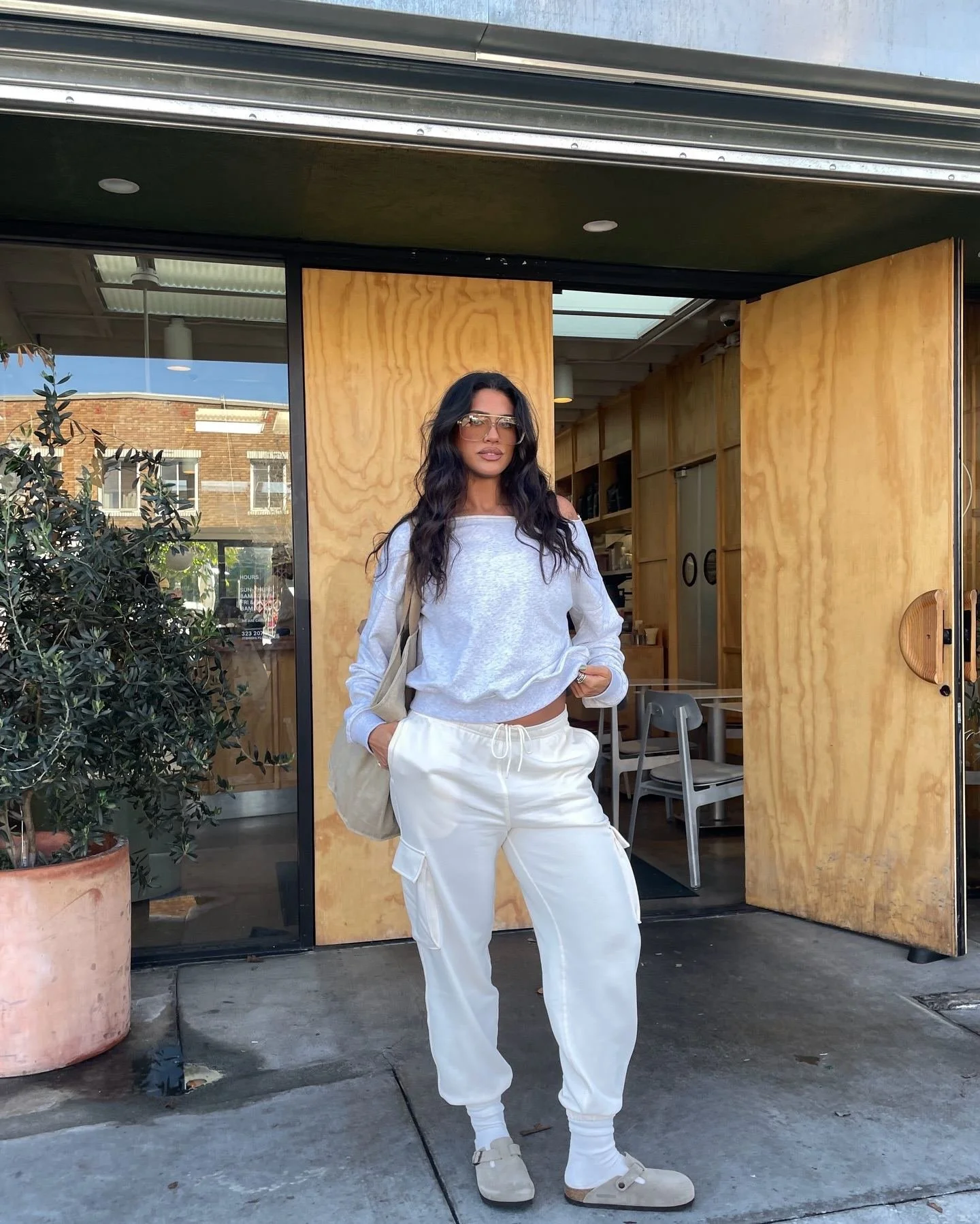 Woman standing outside a cafe doorway wearing sunglasses, a white long sleeve shirt, white cargo pants, and beige sneakers, with a beige bag over her shoulder.