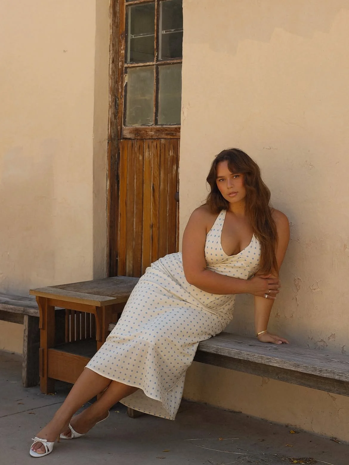 A woman in a white polka dot dress sitting on a wooden bench against a textured beige wall, with an old wooden window partly in view.