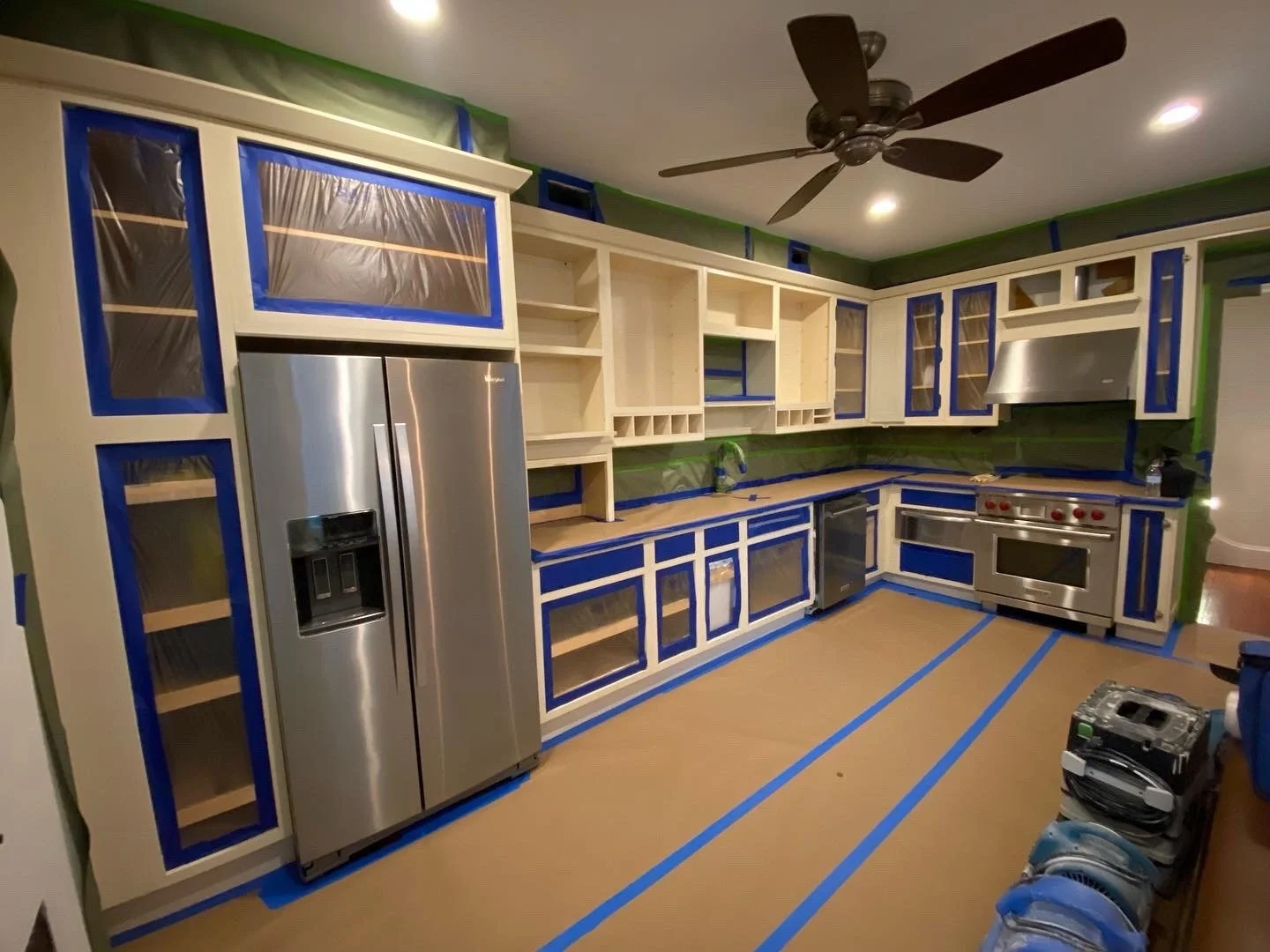 Kitchen under renovation with cabinets taped off in blue painter's tape, stainless steel refrigerator, stove, and dishwasher, green walls, ceiling fan, and construction tools on the floor.