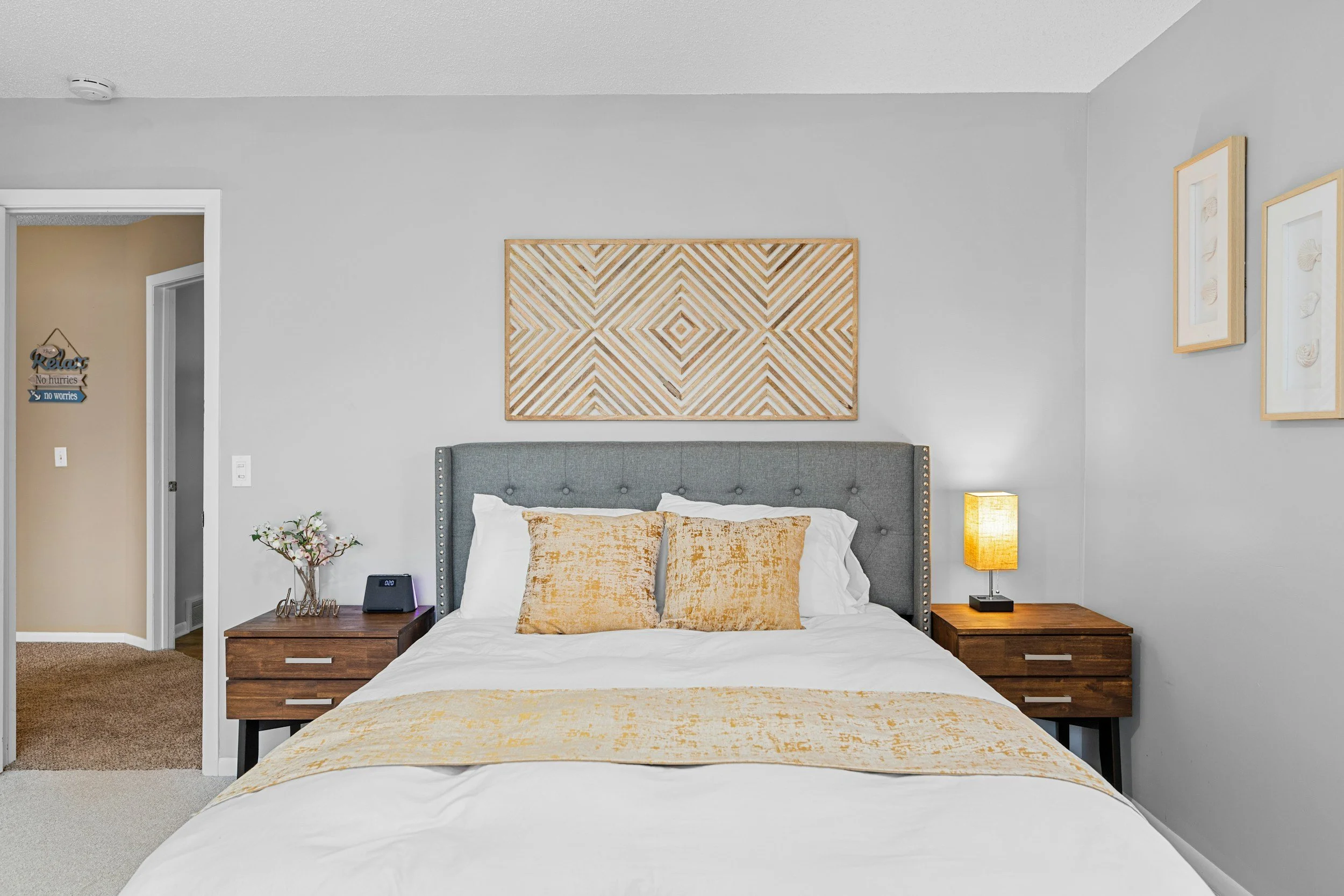 A neatly made bed with a gray upholstered headboard, white bedding, and beige decorative pillows, flanked by wooden nightstands, with a modern box lamp and flowers on one side, and artwork above the bed, in a bright bedroom with light gray walls.
