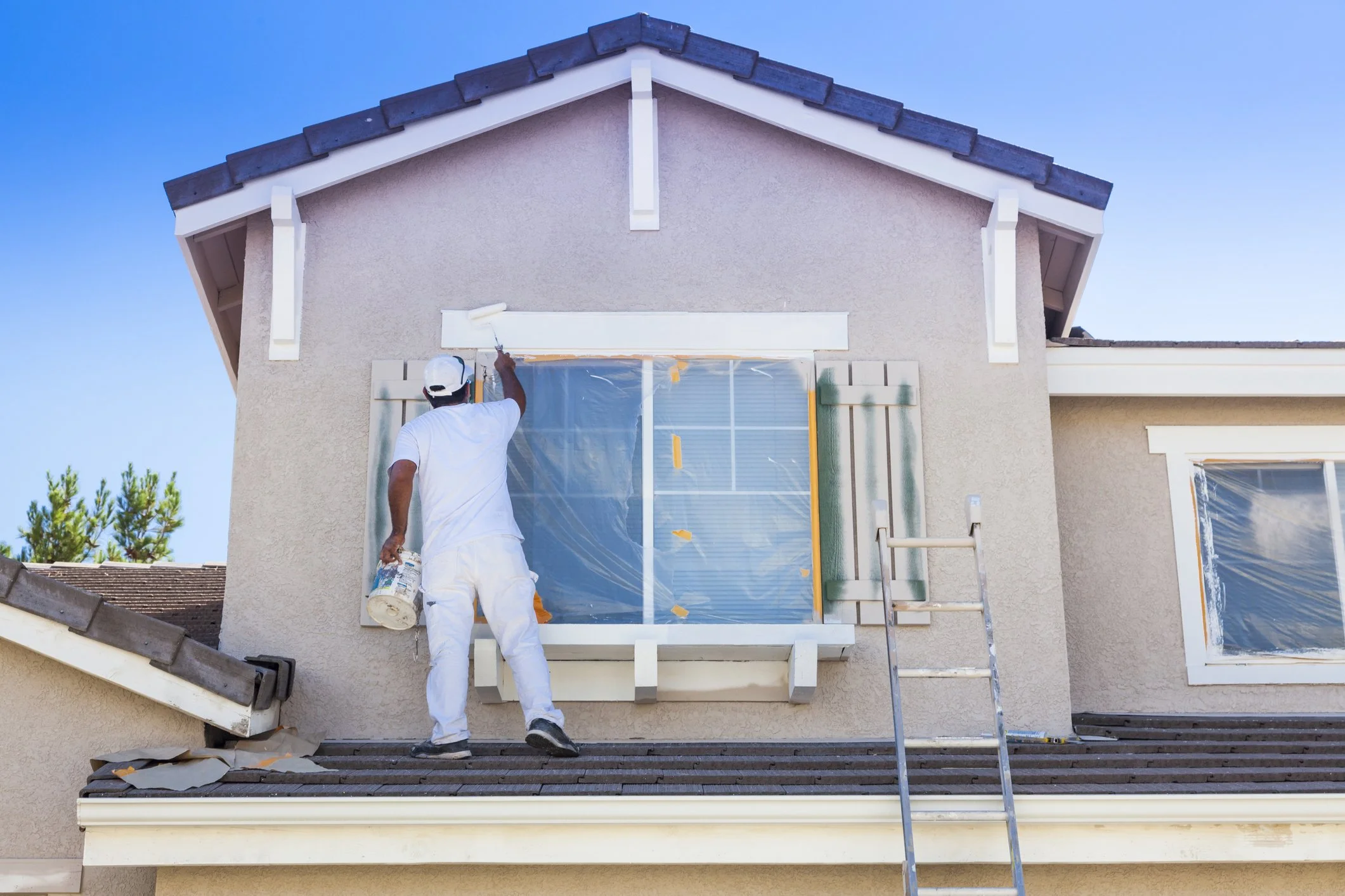 A man painting the trim of a house on a ladder with painter's tape and plastic cloth covering the window, during daytime.