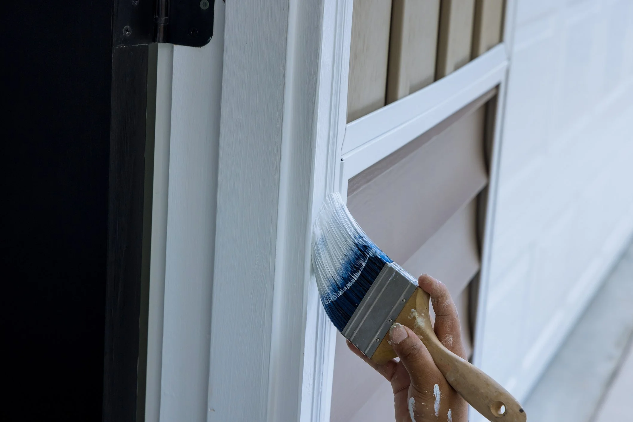 A person is painting the trim of a house with a large paintbrush. The trim is white, and there are beige and brown sections of siding in the background.