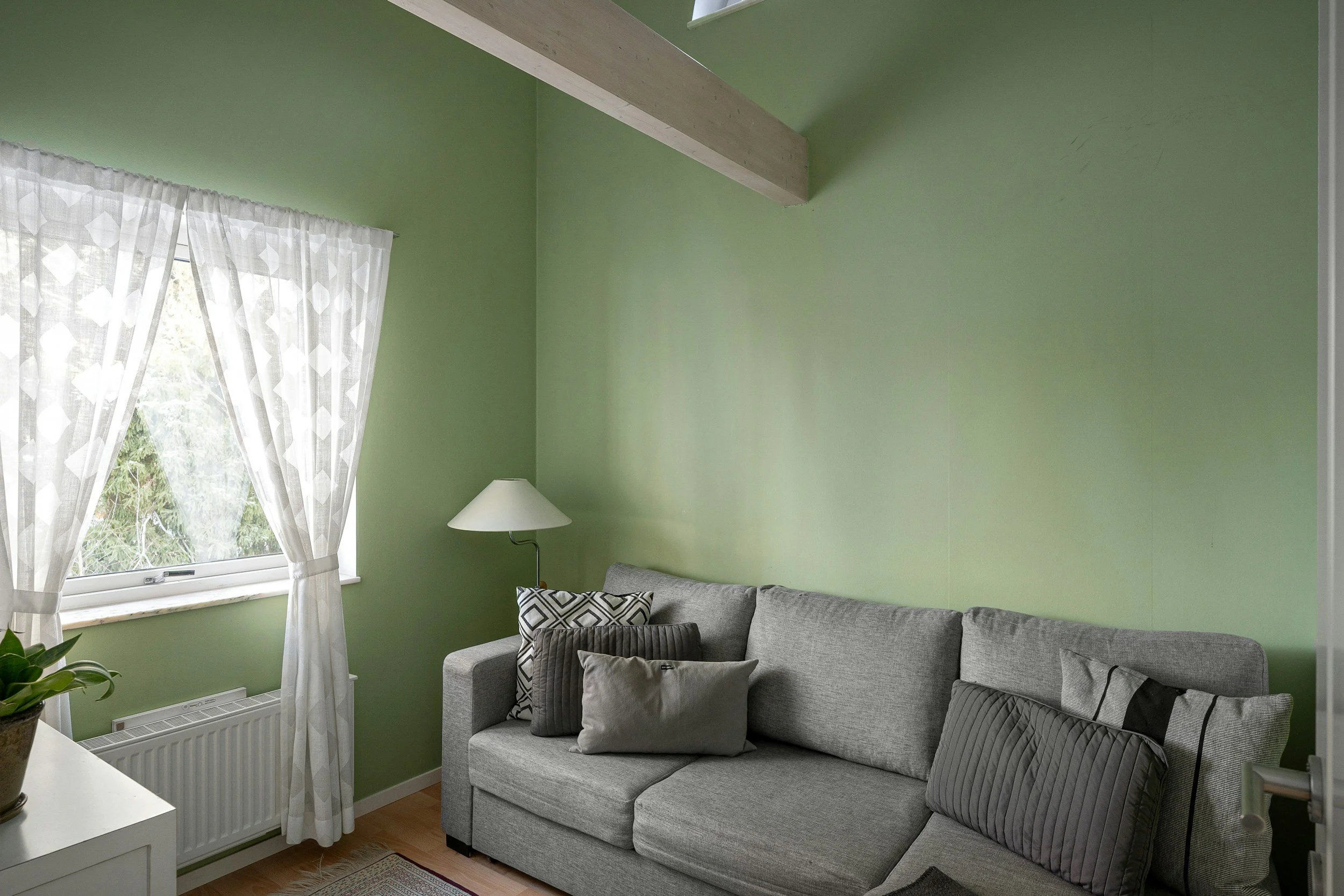 Living room with a gray sofa, pillows, a white lamp, a window with white curtains, and a green wall.