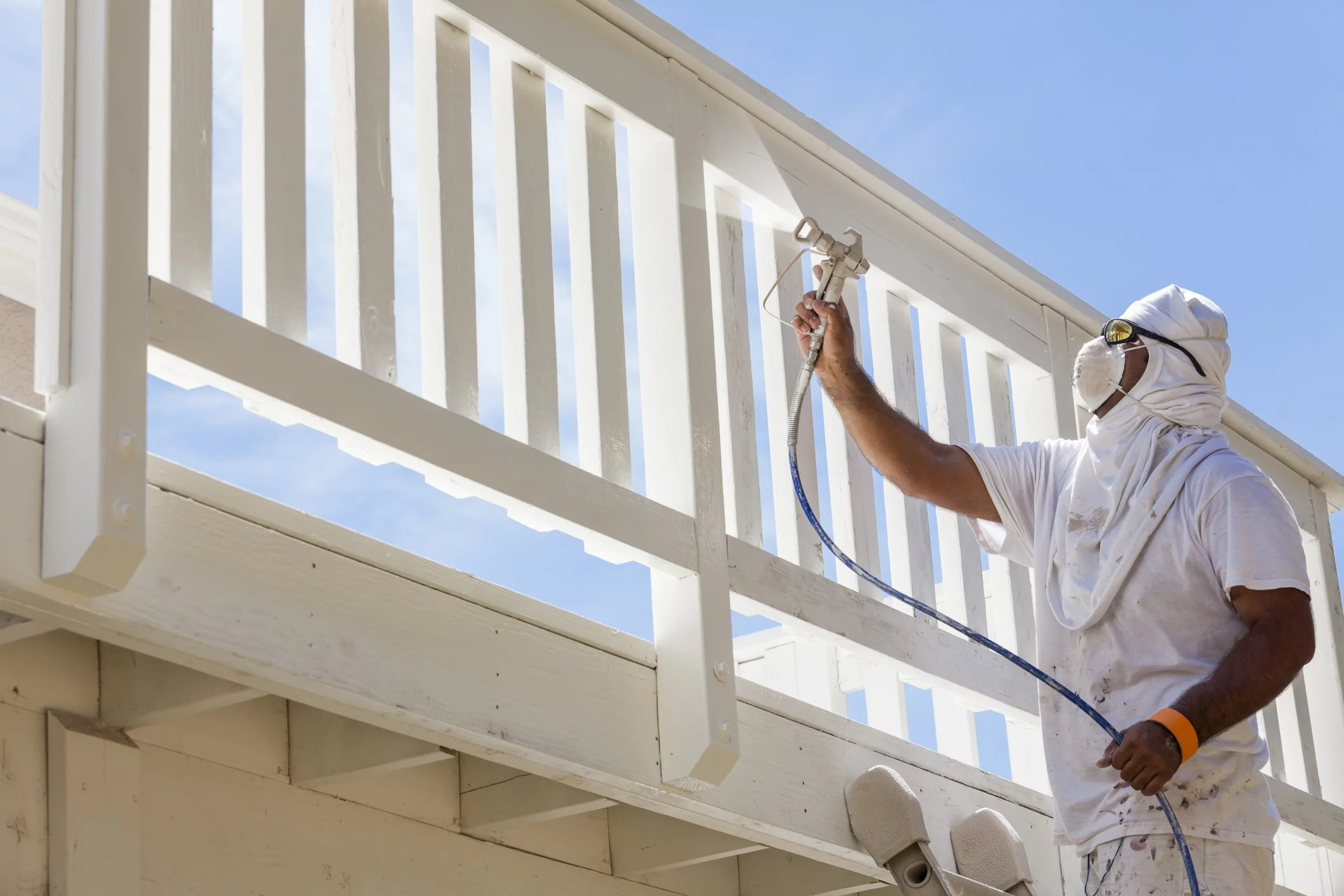 A man painting a white wooden railing on a balcony outdoors on a sunny day, wearing protective goggles, a mask, and white clothes covered in paint splatters, using a spray gun.