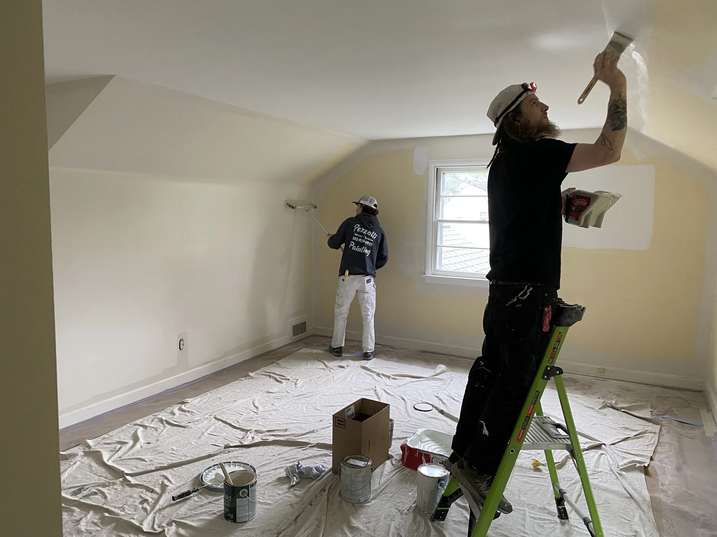 Two men painting the walls of a room. One is on a ladder applying paint with a brush, and the other is using a roller on an extension pole to paint the wall near the window. The room is being prepped for painting with drop cloths on the floor and paint cans nearby.