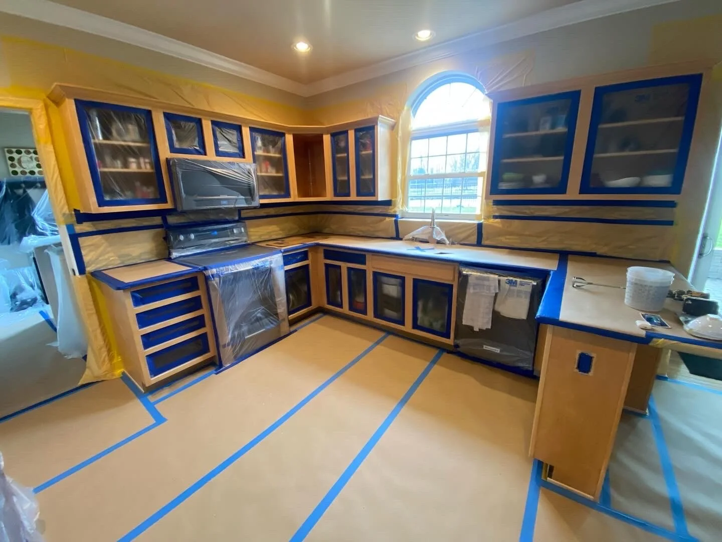 Kitchen under renovation with cabinets covered in protective plastic and painter's tape, appliances also wrapped, and protective paper on the floor.
