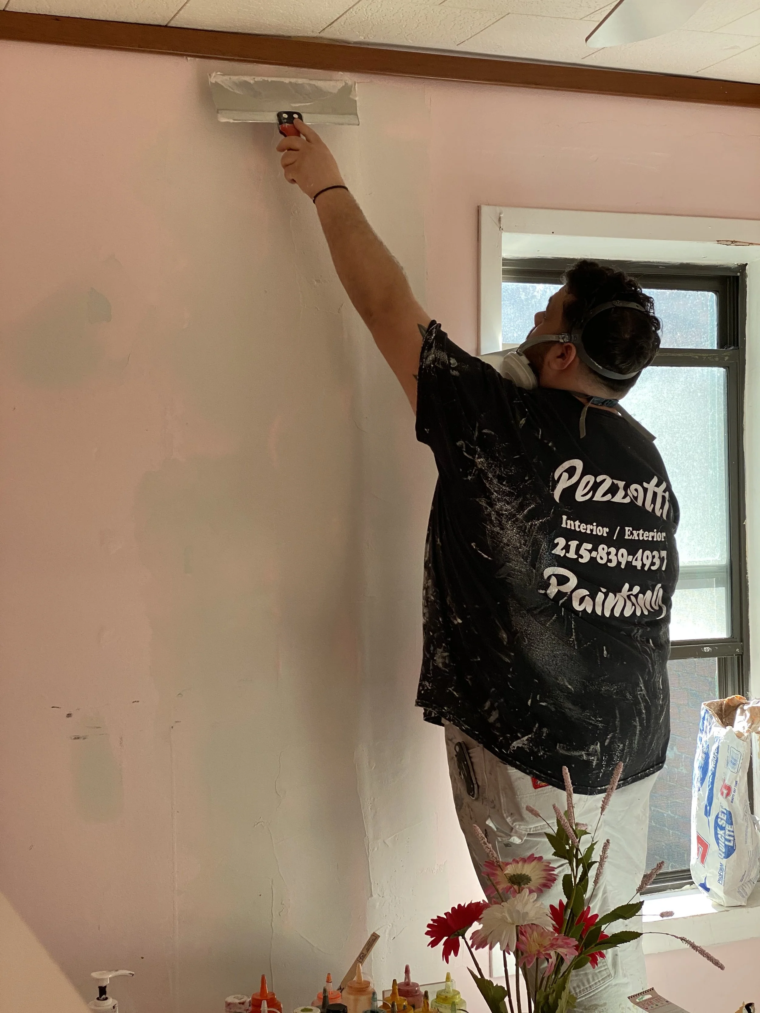 A man painting a wall with a paint roller, wearing a black T-shirt with white text, standing next to a window and a vase of flowers on a table with various paint bottles.