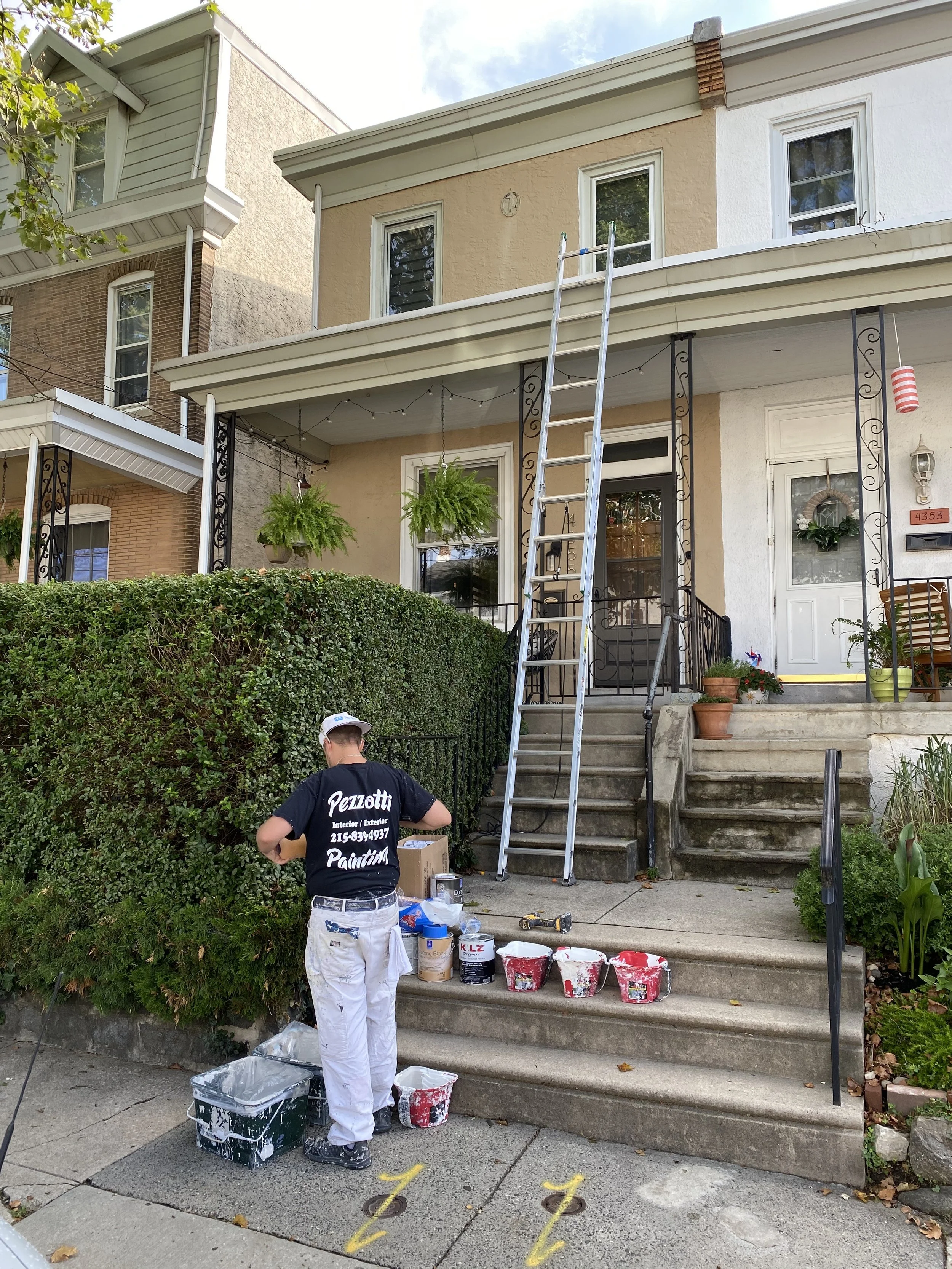 Man painting the front porch stairs of a house with buckets of paint and painting supplies around him. The house has a front door, potted plants, hanging planters, and a ladder leaning against the second story of the house.