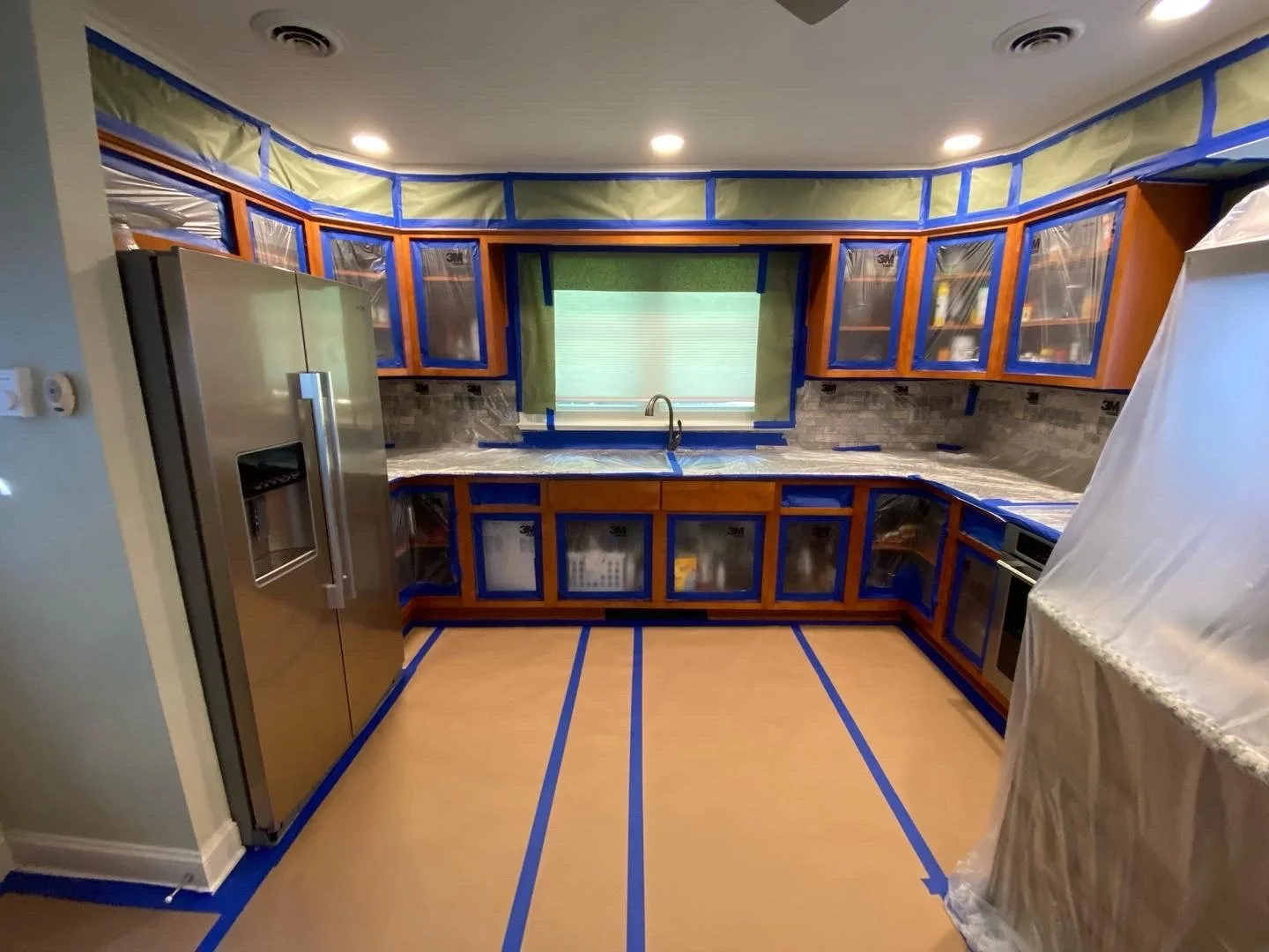 A kitchen under renovation with painter's tape on the floor, cabinetry, a stainless steel refrigerator, and a window above the sink covered in green tape, indicating ongoing updates.