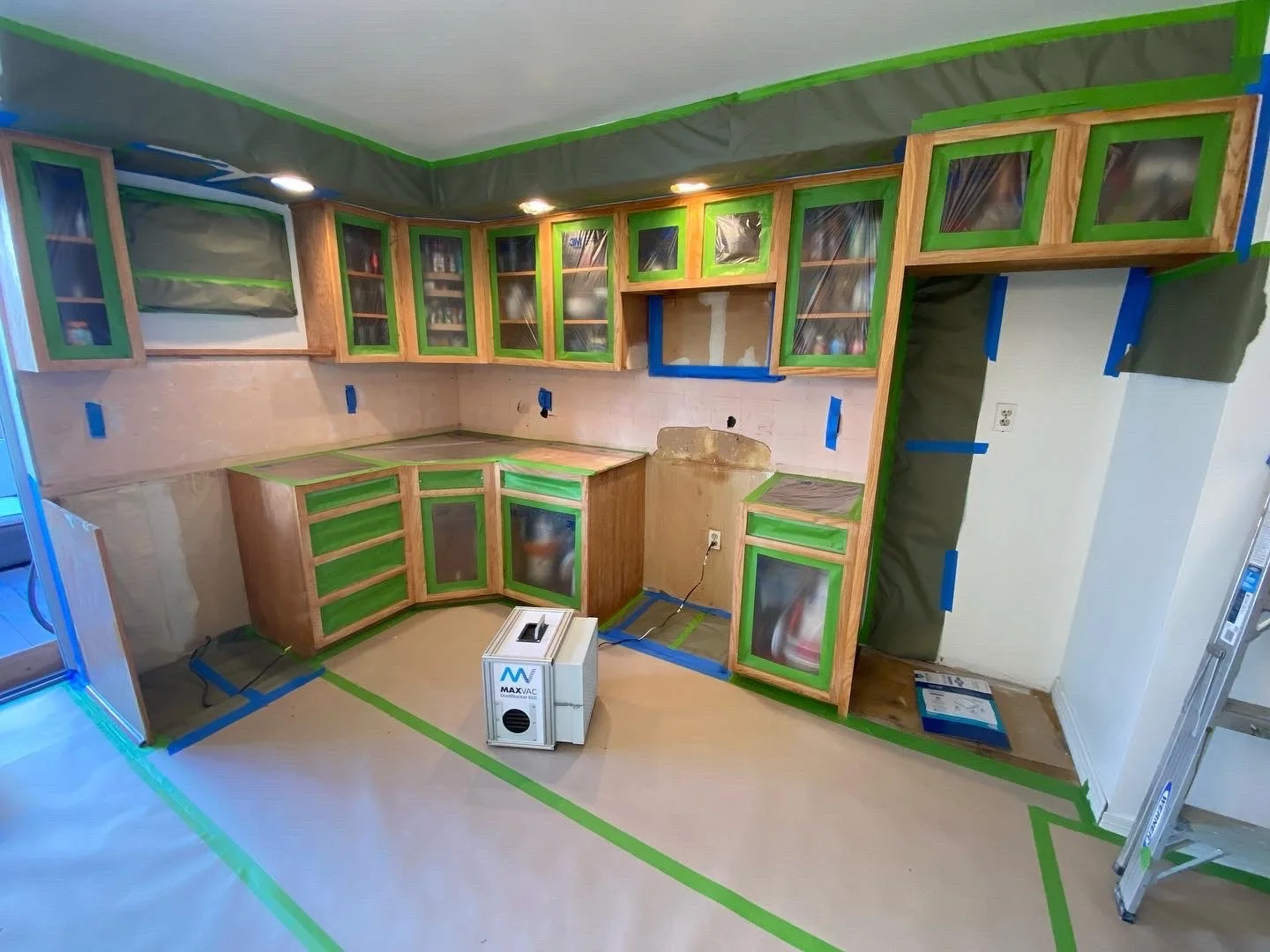 Kitchen under renovation with unfinished cabinets, covered in green painter's tape, a ladder to the right, a portable air purifier on the floor, and protective paper covering the floor.