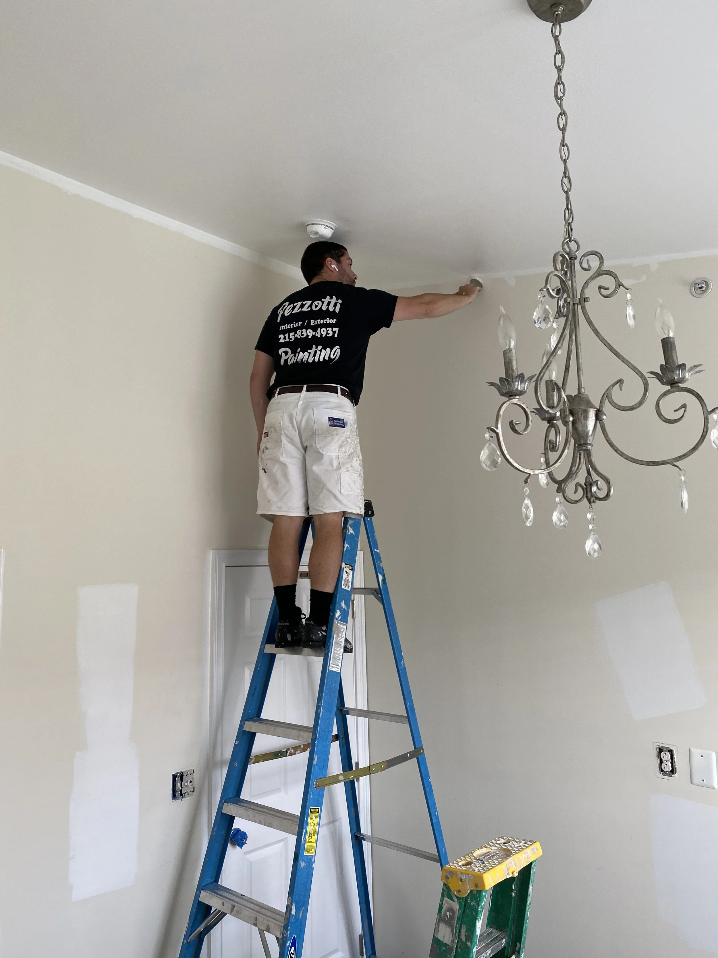 A man standing on a ladder painting the ceiling of a room, with a chandelier hanging nearby.