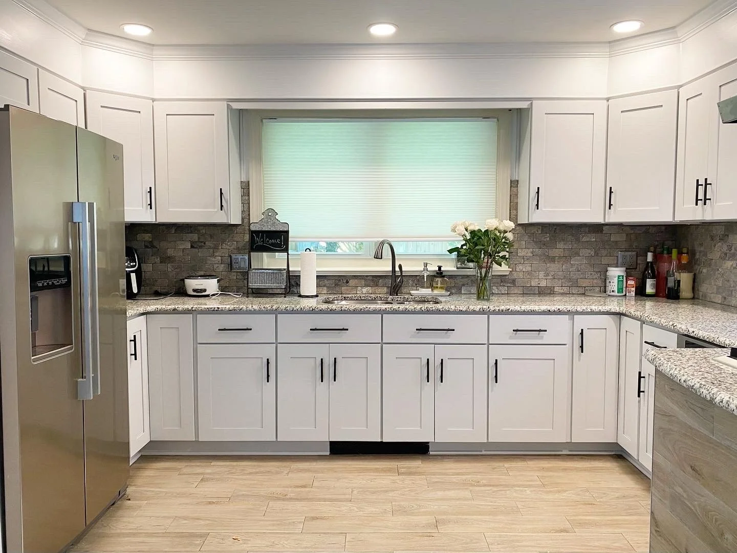 Modern kitchen with white cabinets, granite countertops, a stainless steel refrigerator, and a window with a green blind, decorated with a vase of white flowers and various small kitchen items.