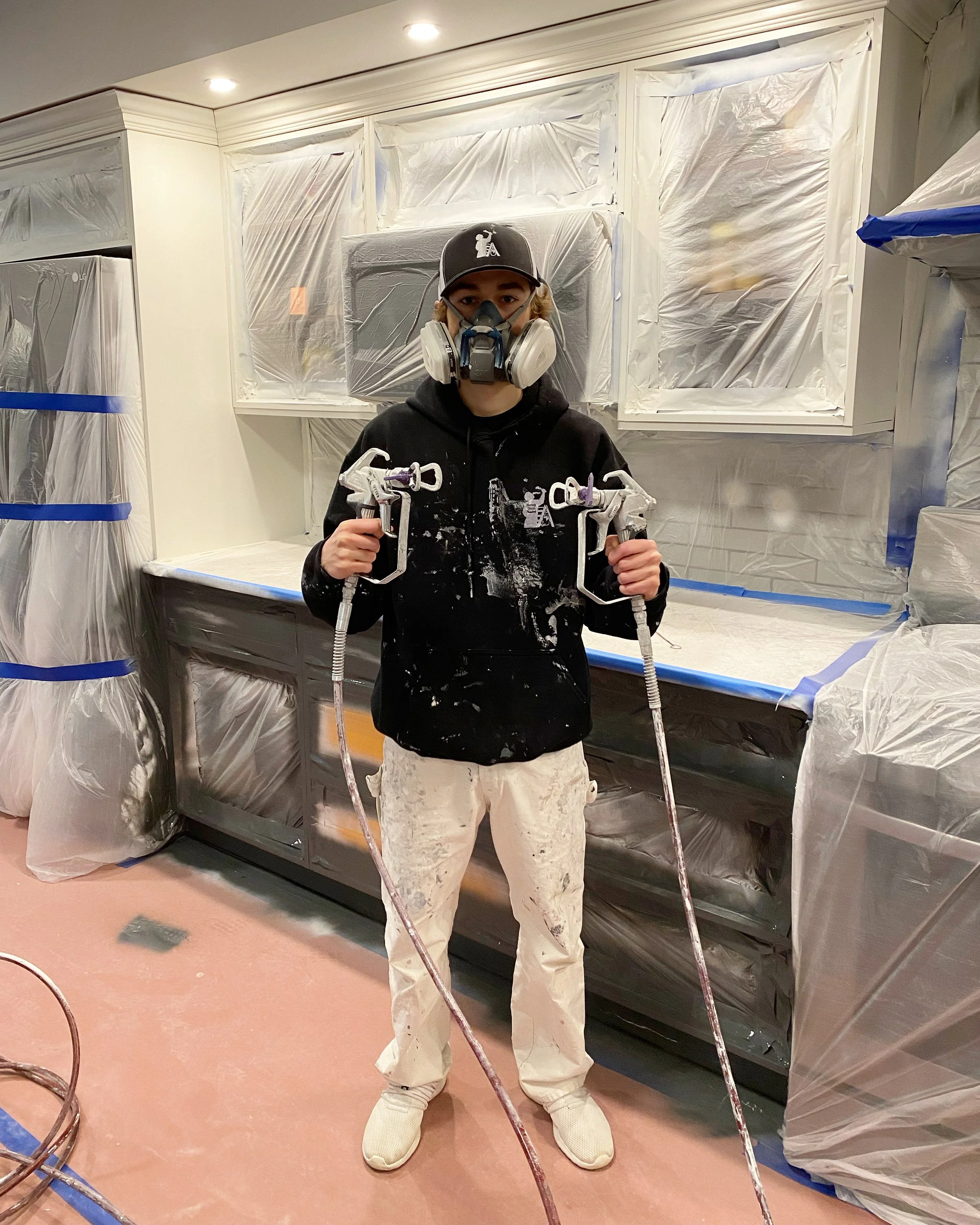 A person wearing a respirator mask, black hoodie, white paint-stained pants, and a baseball cap, holding paint spray guns, standing in a room under renovation with plastic sheeting covering the walls and cabinets.
