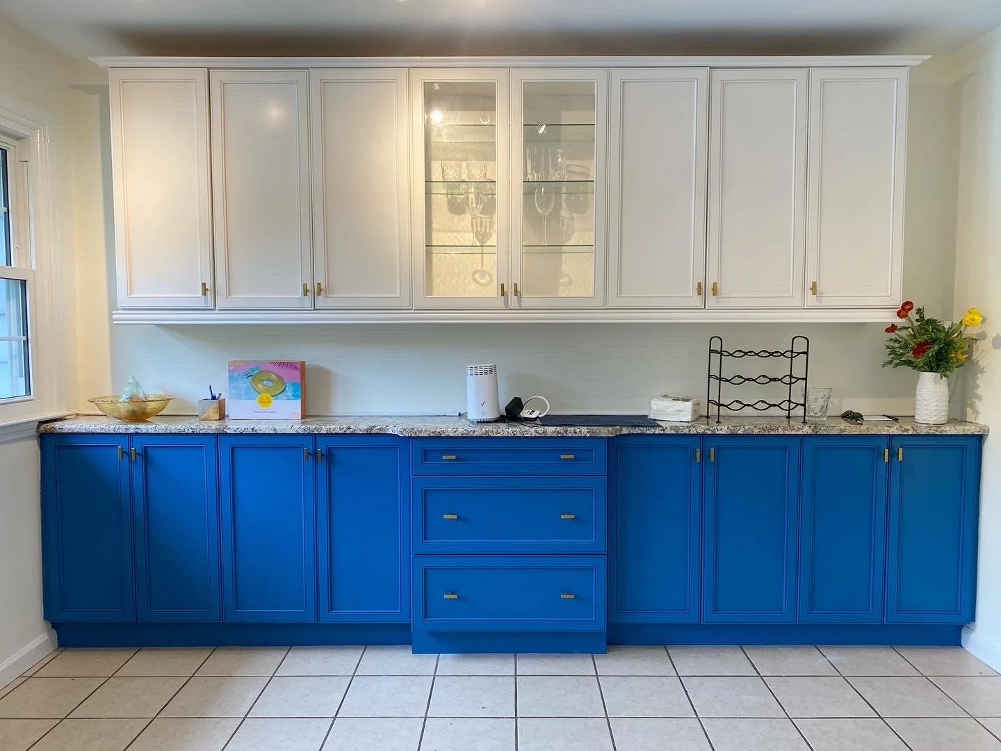 Kitchen with white upper cabinets, blue lower cabinets, granite countertop, and tile flooring. Items include a bowl, a small picture, a diffuser, a dish rack, a vase with flowers, and some small objects.