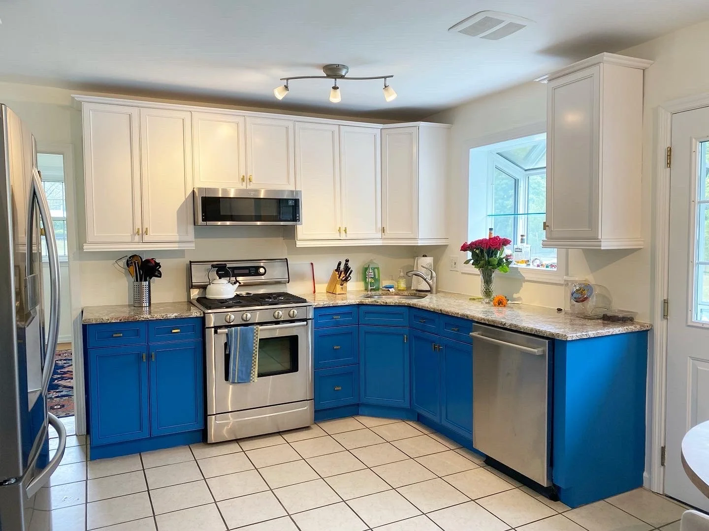 A kitchen with white upper cabinets, blue lower cabinets, a stainless steel stove and microwave, a granite countertop, a dishwasher, and a window with a vase of pink flowers on the counter.