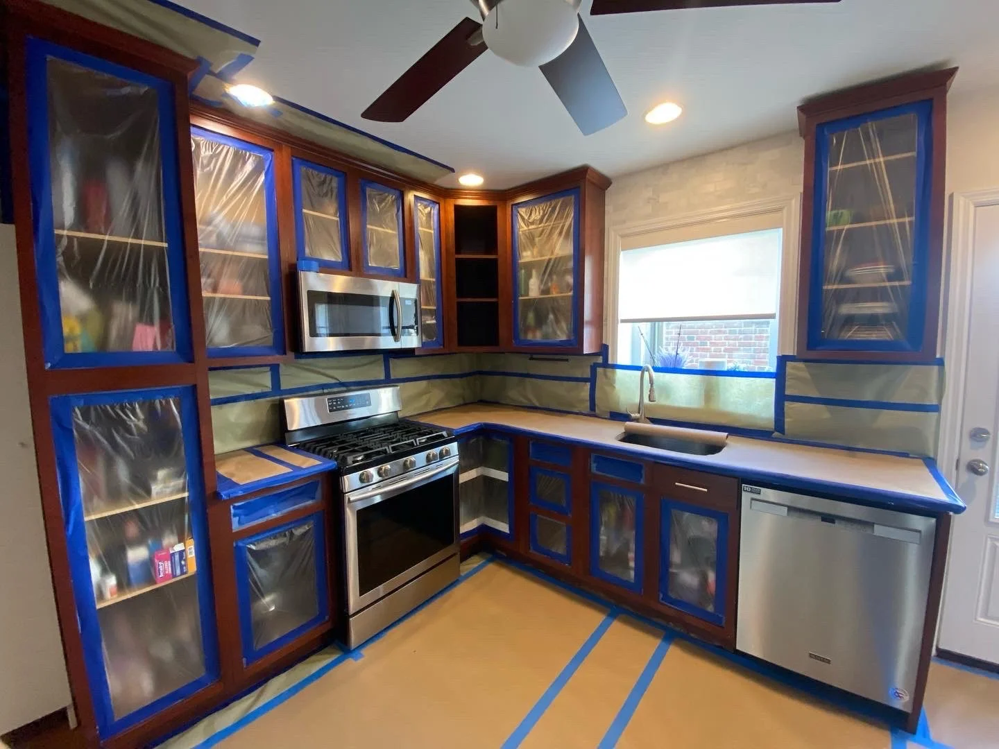 Kitchen being renovated with cabinets covered in plastic and painter's tape, stove, microwave, dishwasher, sink, window, ceiling fan, and recessed lights.
