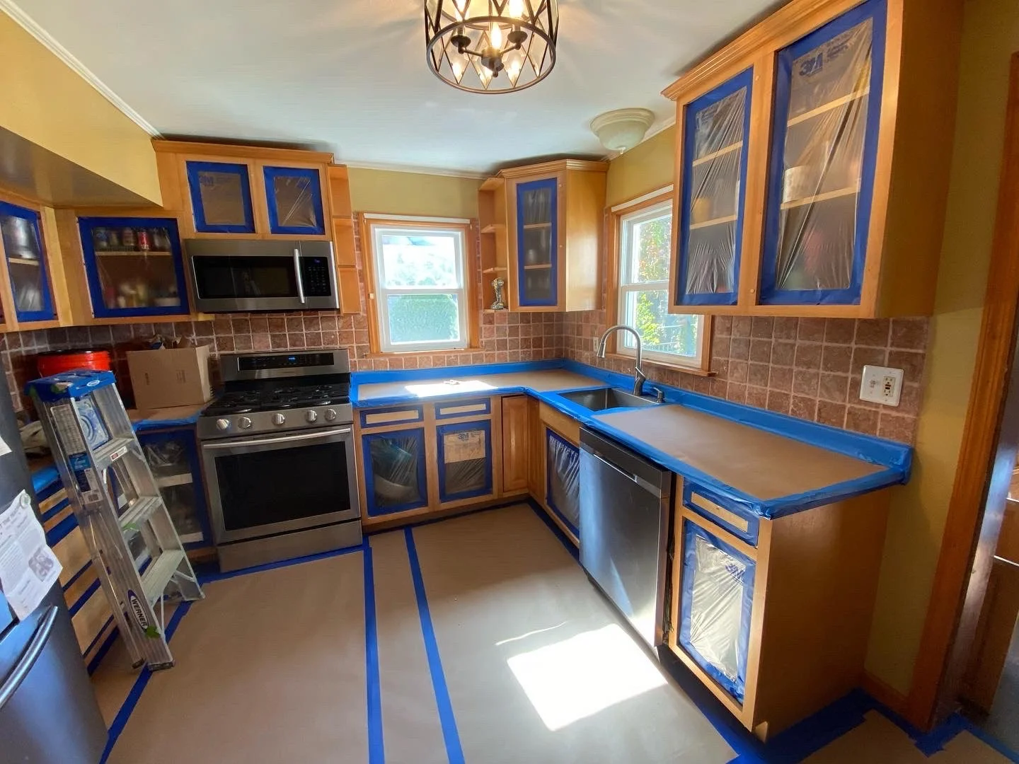 Kitchen in the process of renovation with blue painter's tape covering cabinet edges and countertops, natural light coming through windows, and stainless steel appliances.