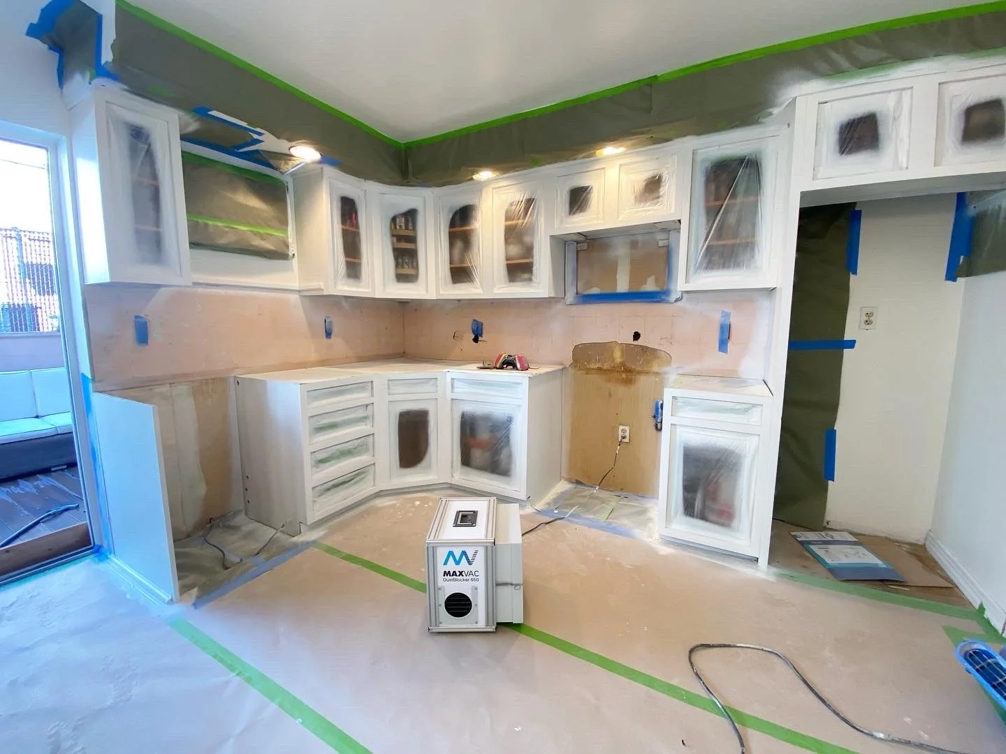 Kitchen under construction with white cabinets covered in protective plastic, green painter's tape on the ceiling and floor, and a portable air purifier in the center.