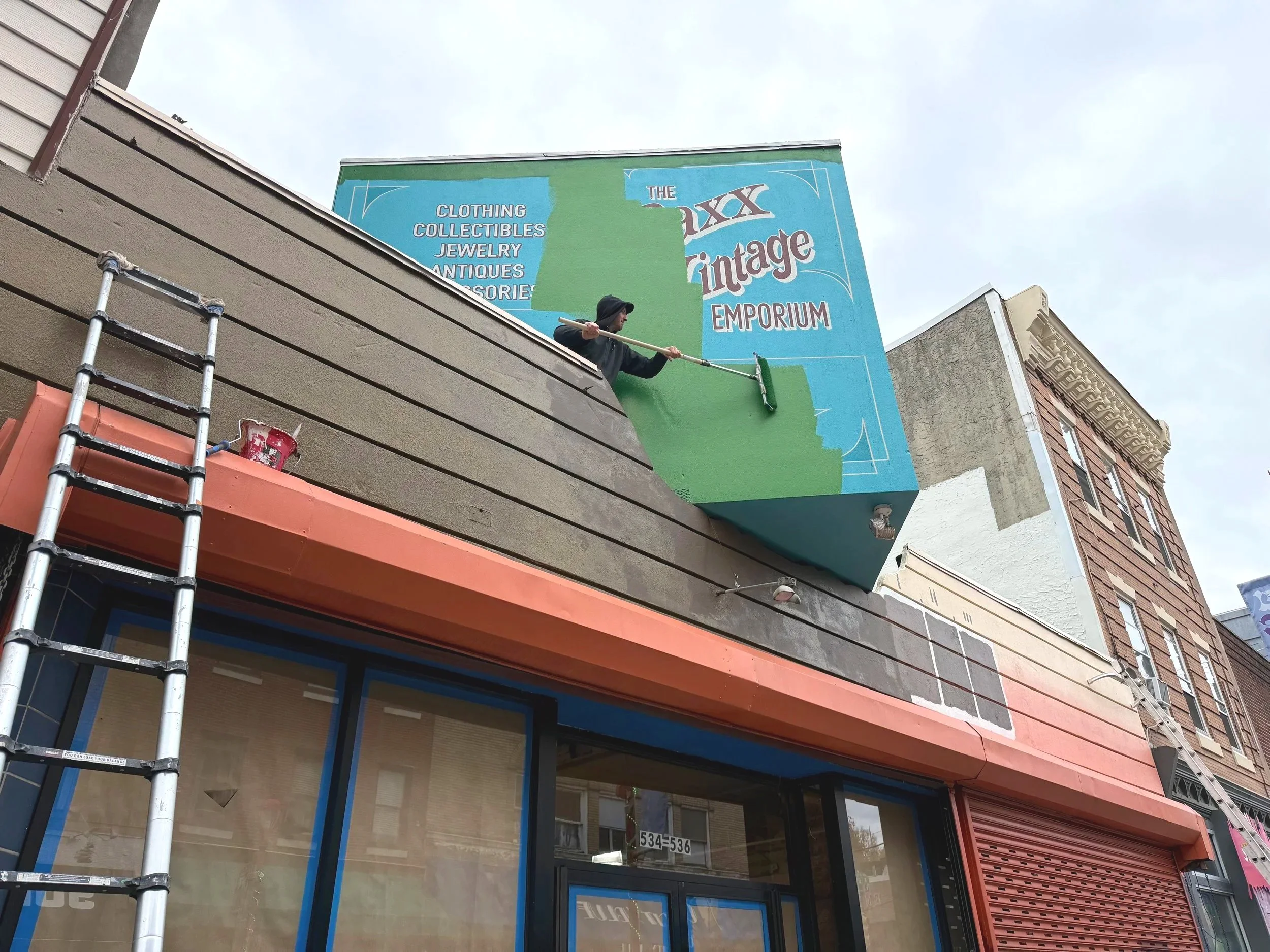 A person painting a large blue and green sign on the exterior of a building with a paint roller. The sign reads "The XXX Vintage Emporium" and lists items like clothing, collectibles, jewelry, antiques, and more. There is a ladder propped against the building and a red bucket of paint next to it.